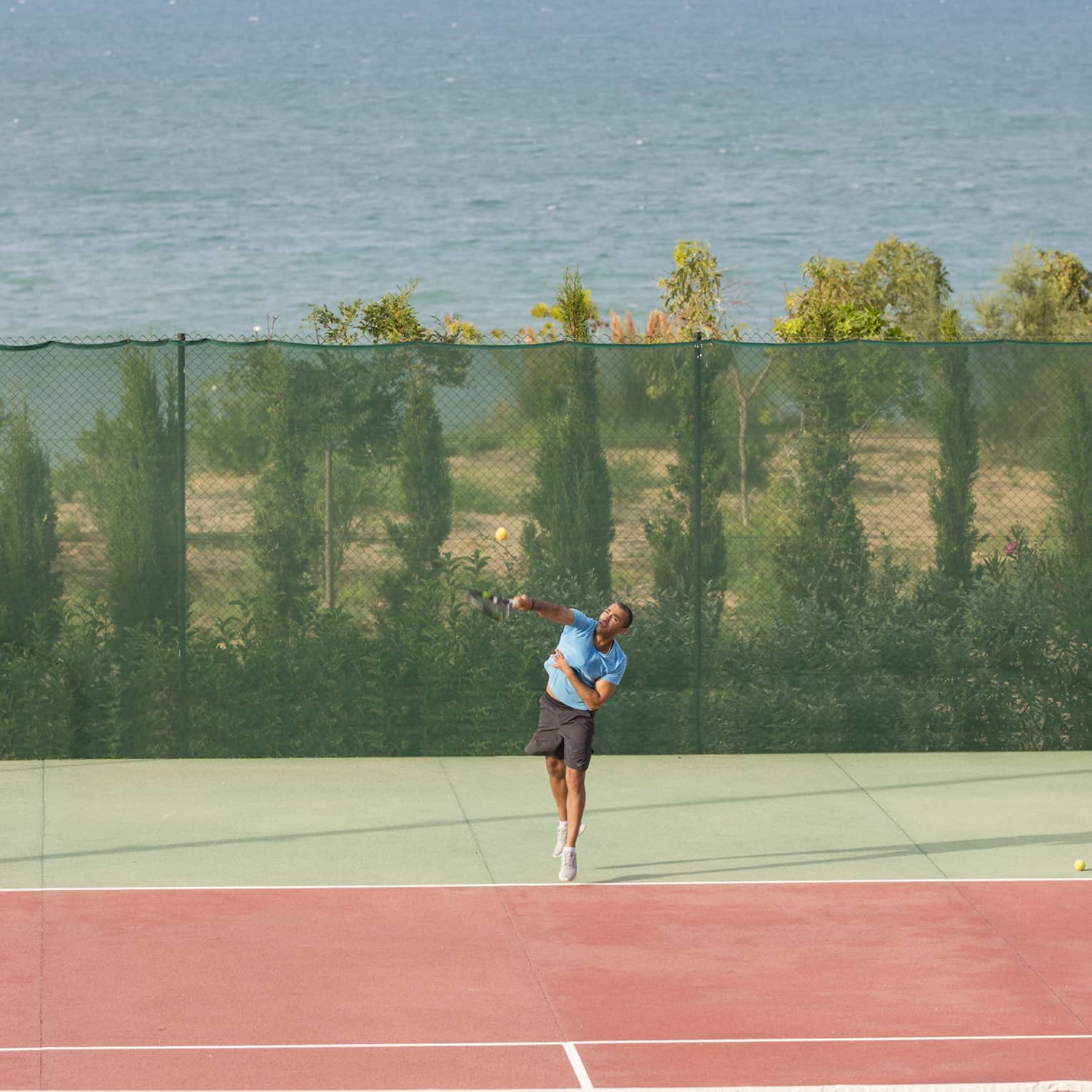 Man in blue shirt and black shorts serving a tennis ball on a seafront tennis court