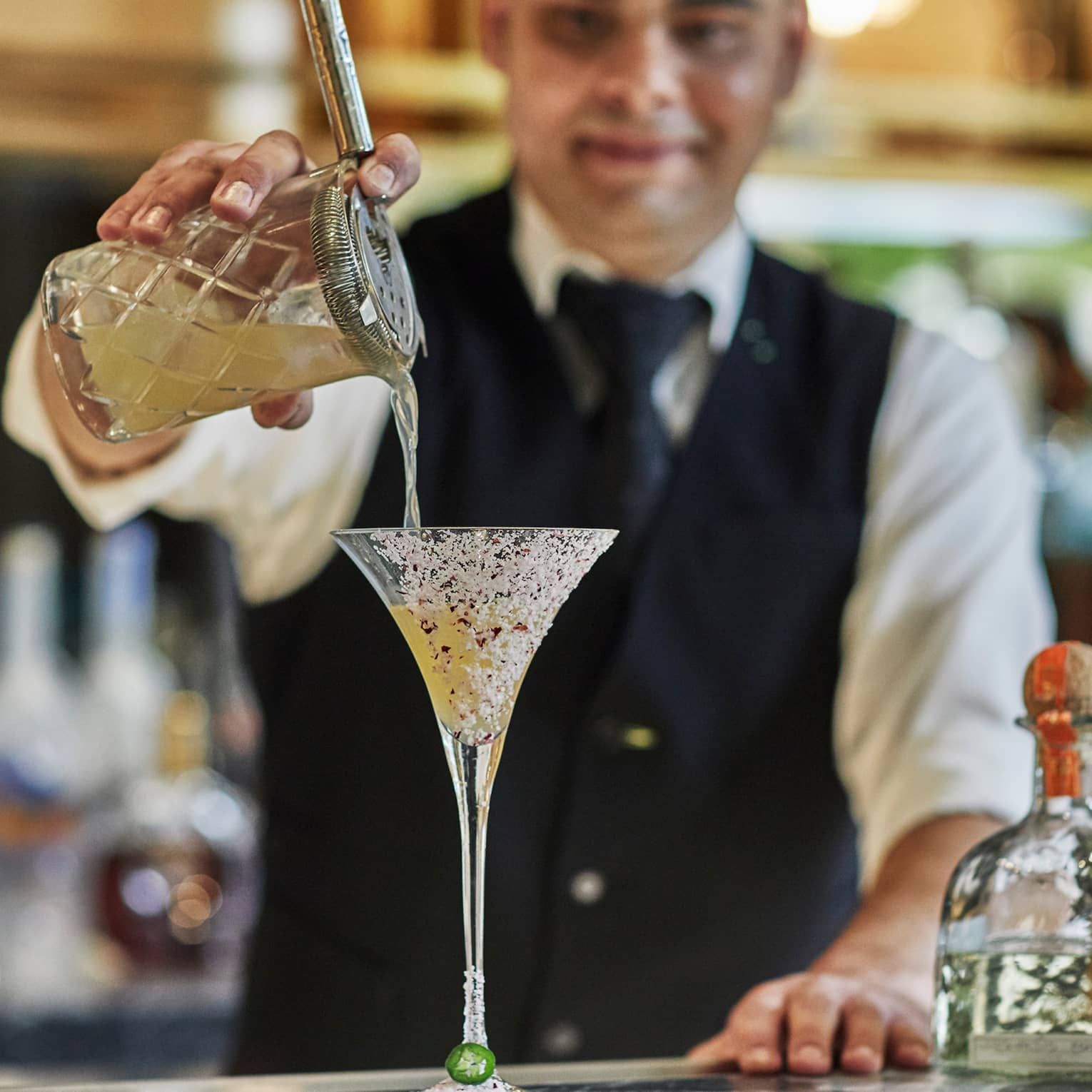 Bartender wearing black vest pours cocktail from crystal shaker into martini glass