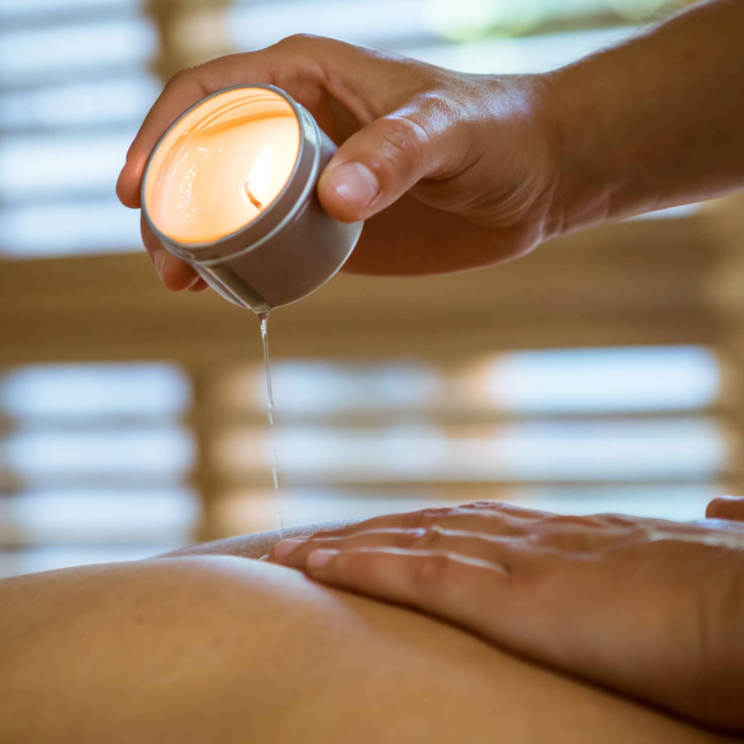 Massage therapist pours hot wax from tealight candle over person's bare back in spa