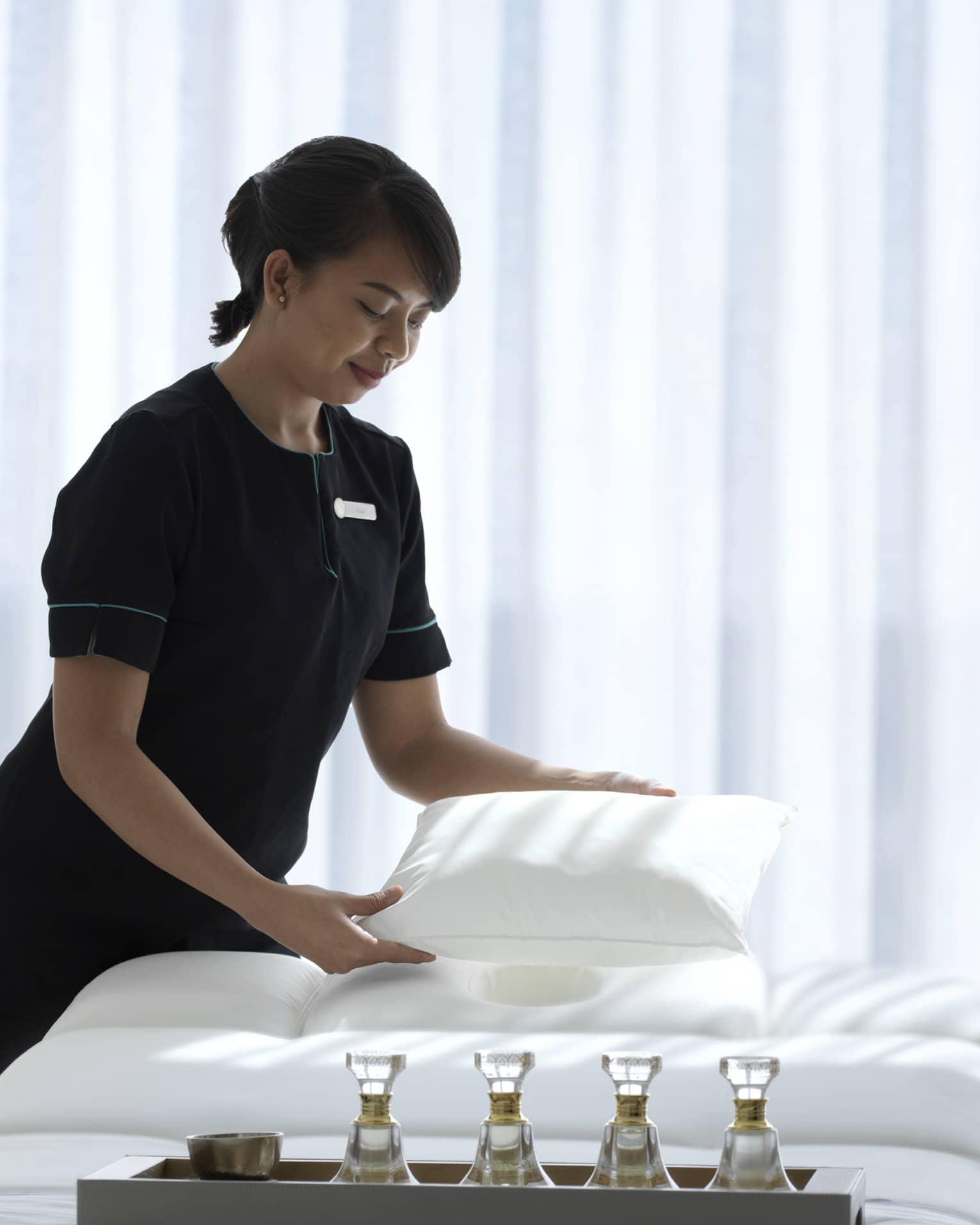 Spa attendant wearing a black uniform fluffs a while pillow on a treatment table covered with all-white linens and a tray filled with four small glass bottles