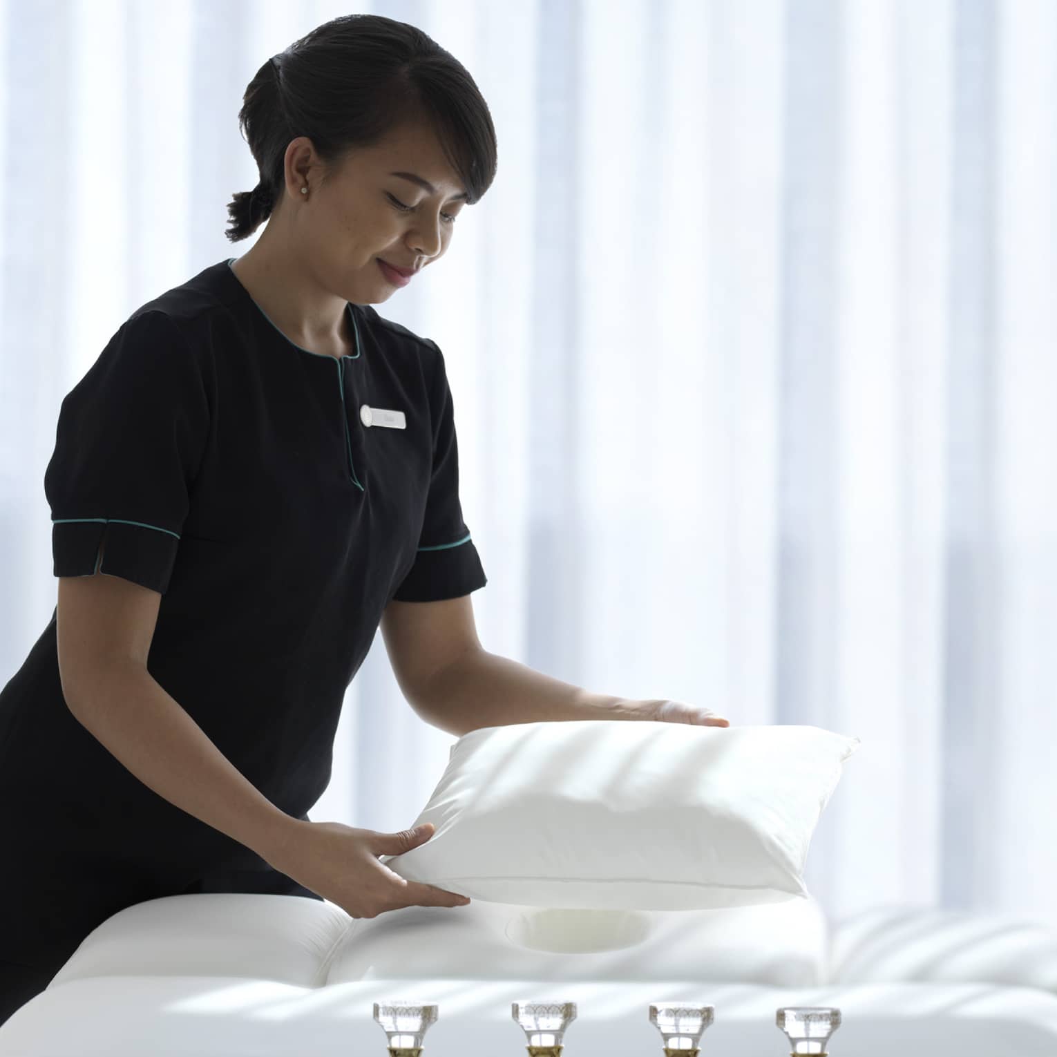 Spa attendant wearing a black uniform fluffs a while pillow on a treatment table covered with all-white linens and a tray filled with four small glass bottles