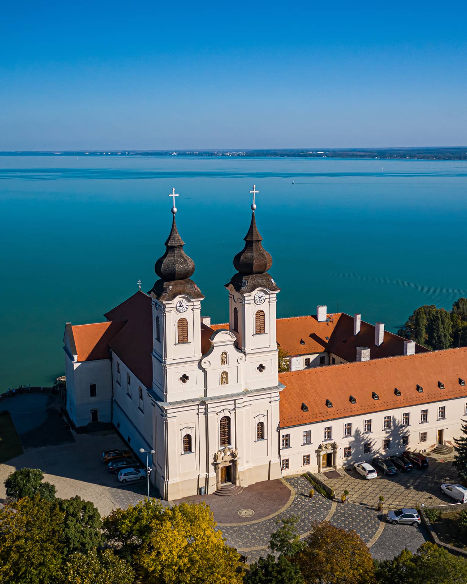 A Benedictine monastery, with white walls and a red tiled roof, sitting beside a large blue lake and surrounded by trees.
