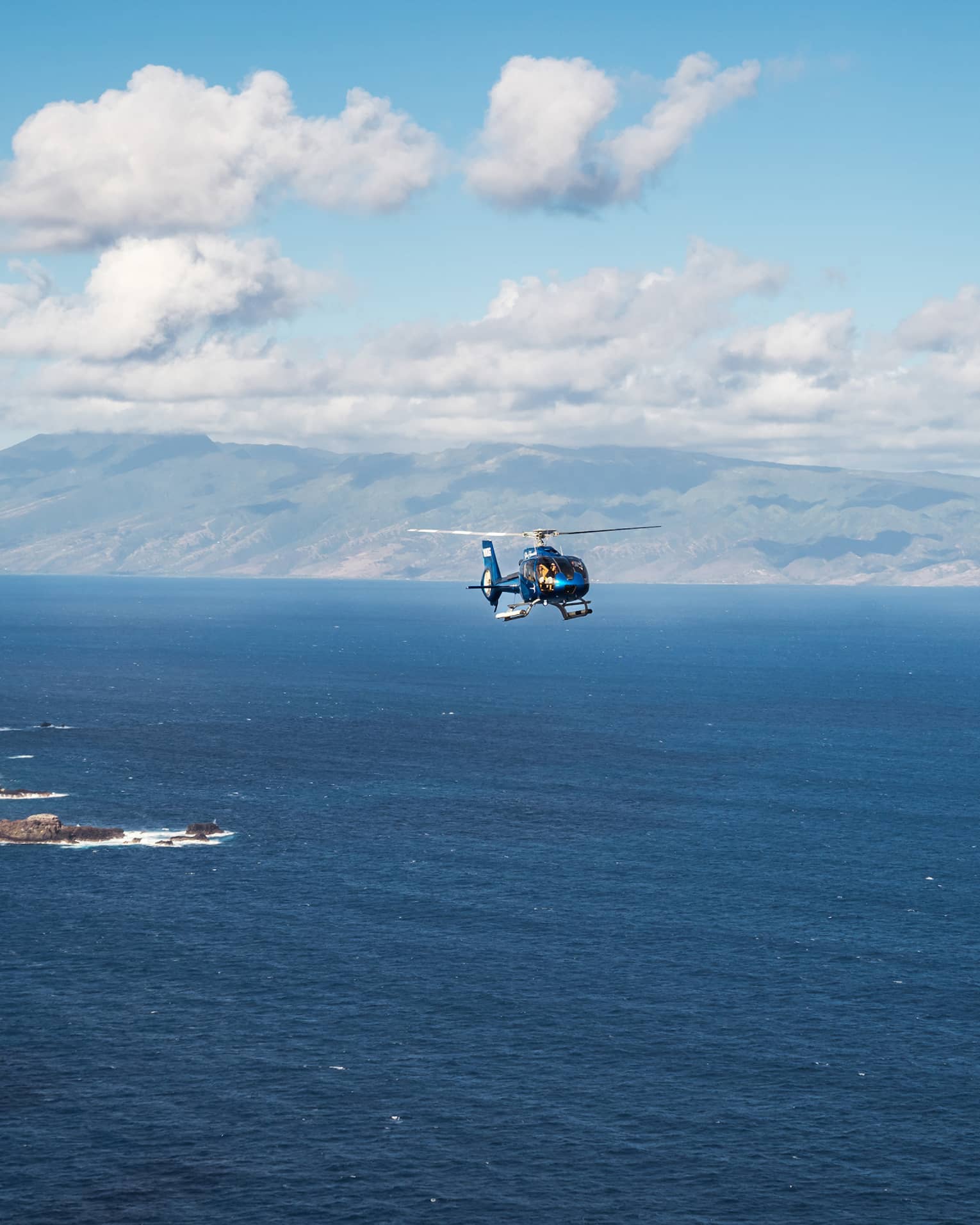 A helicopter flies over open ocean with a mountainous island and blue sky dotted with clouds in the background