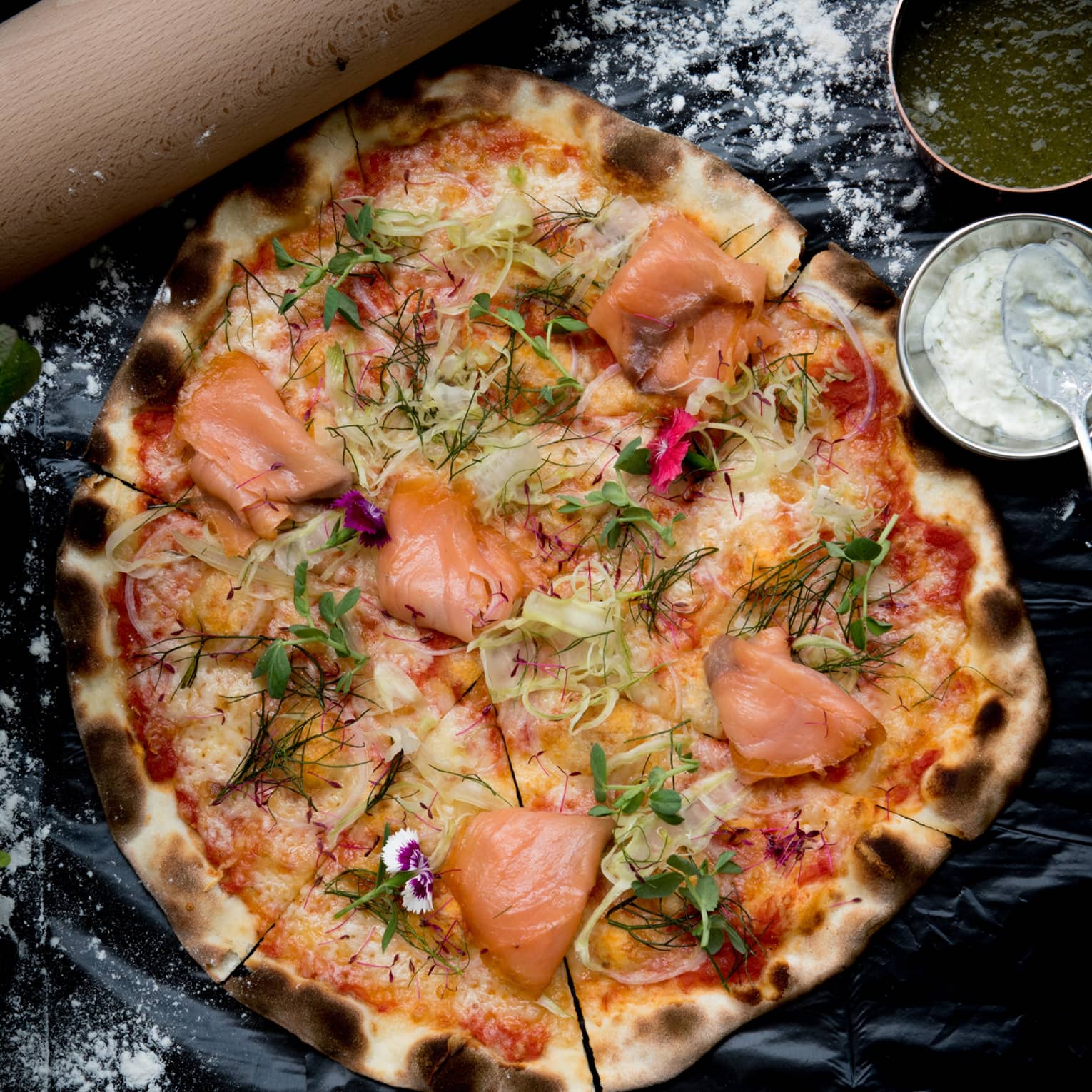 A wood-fired pizza topped with smoked salmon, fennel and dill, bowls of green and white sauces on the side. 