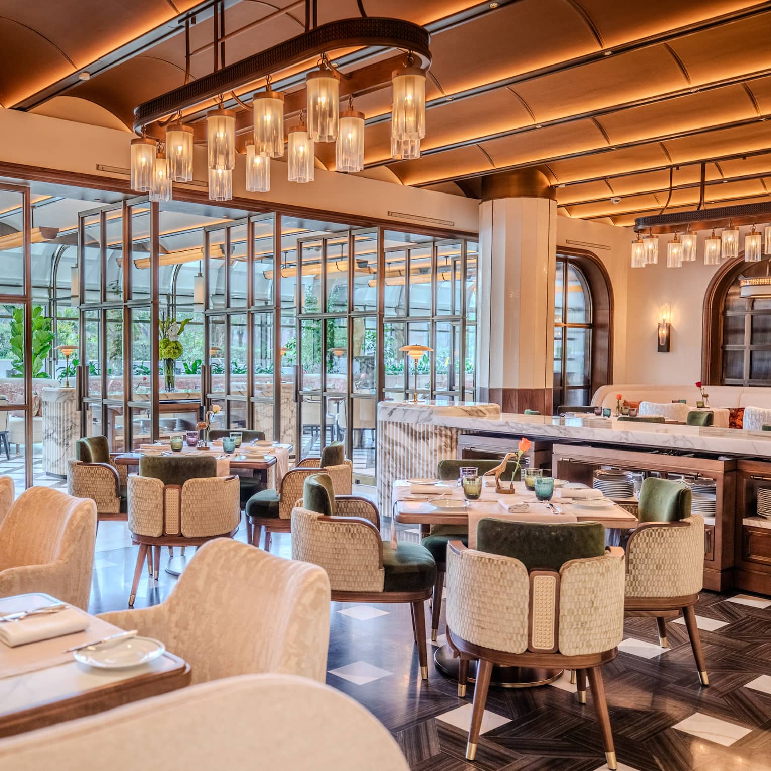 Restaurant dining room featuring tall beamed ceilings, white marble tables, dark flooring and green-and-white velvet chairs all lit by warm overhead lights
