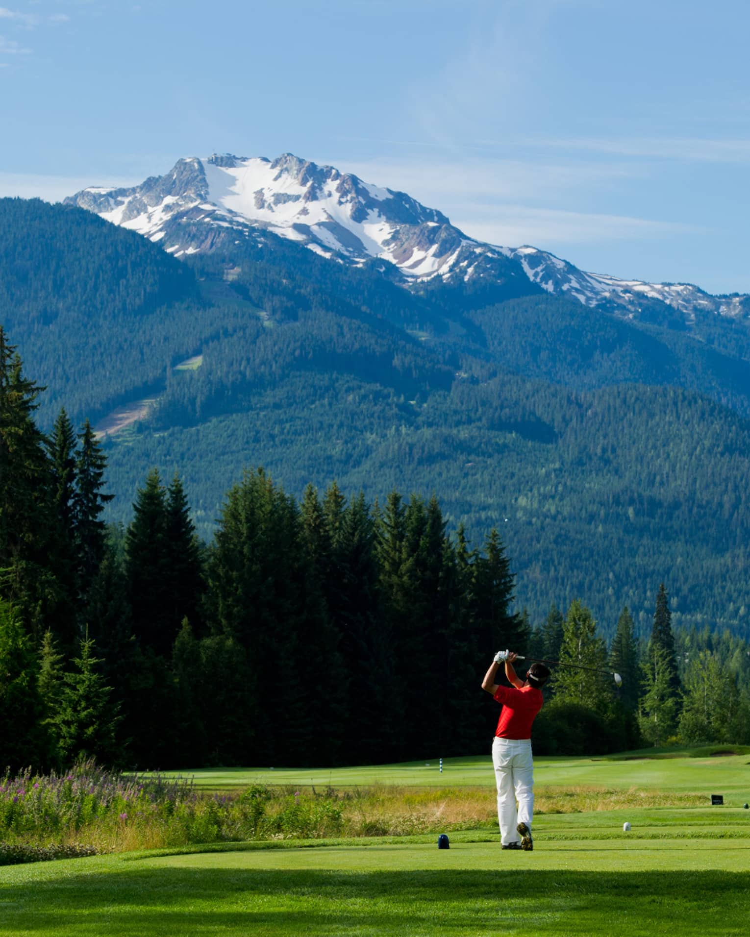 Man wearing red golf shirt swings club on green golf course surrounded by snow-capped mountains