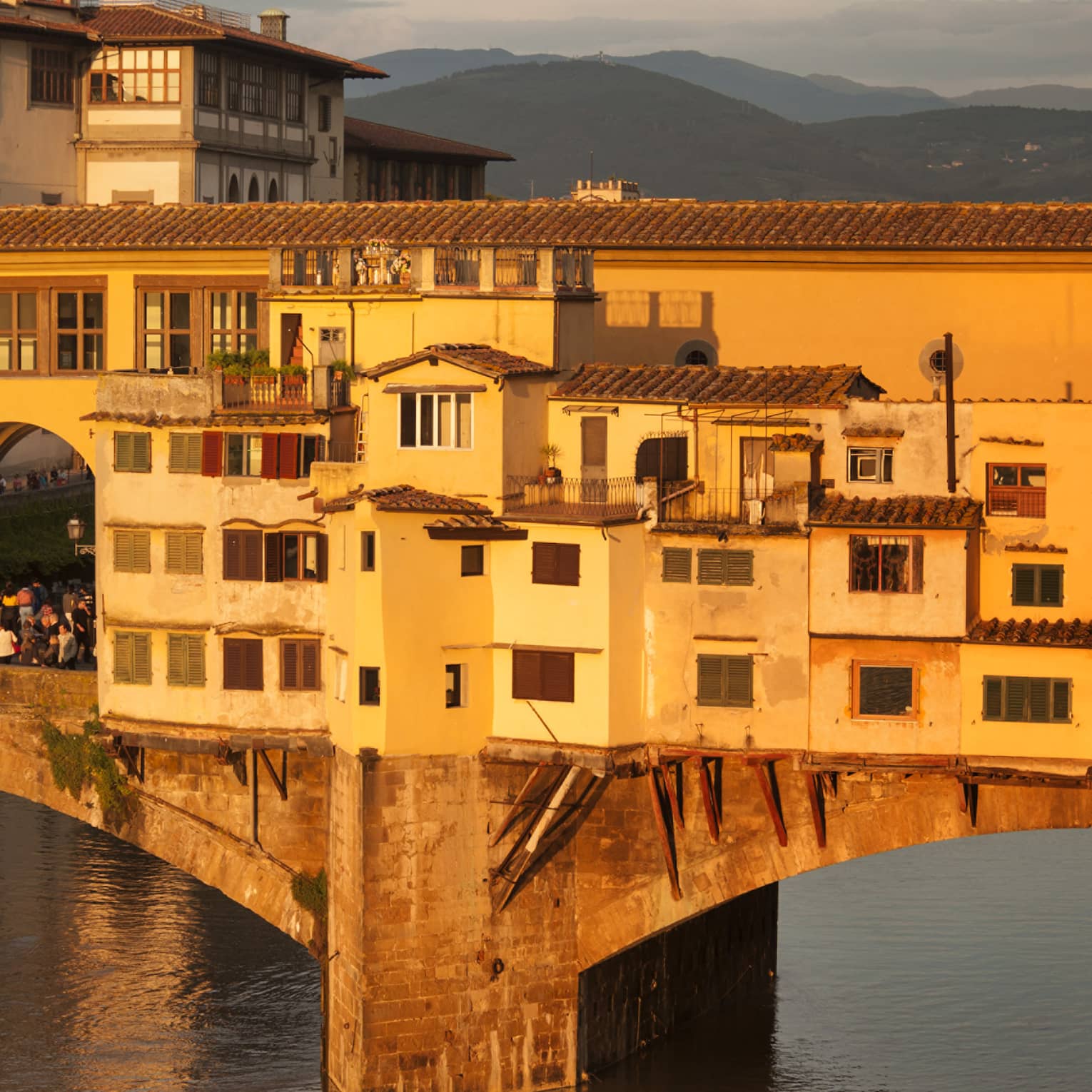 Historic stone bridge with yellow buildings and arched passageways over a river, illuminated by warm sunlight, with mountains visible in the background.