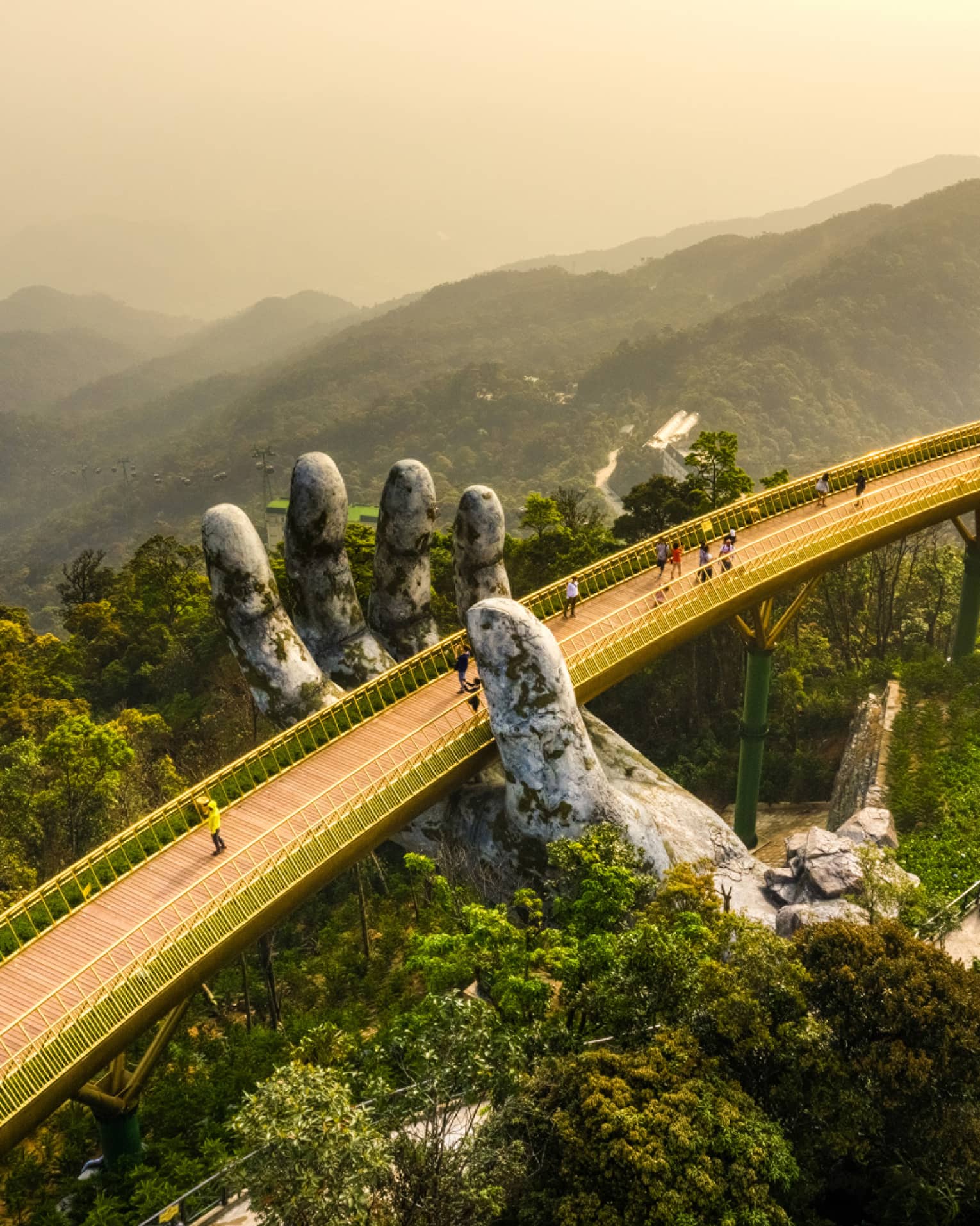 Aerial view of people on a wood-slat bridge above a forest canopy, with golden railings supported by two giant stone hands.