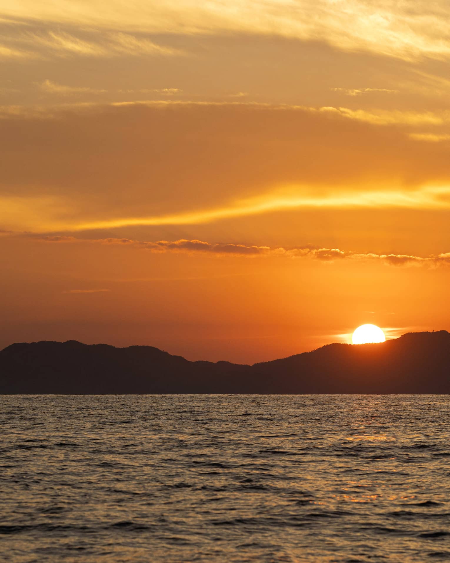 Beyond gently rippling waves, the blazing sun sets behind a hilly island, wispy clouds in the sky awash in its orange glow.