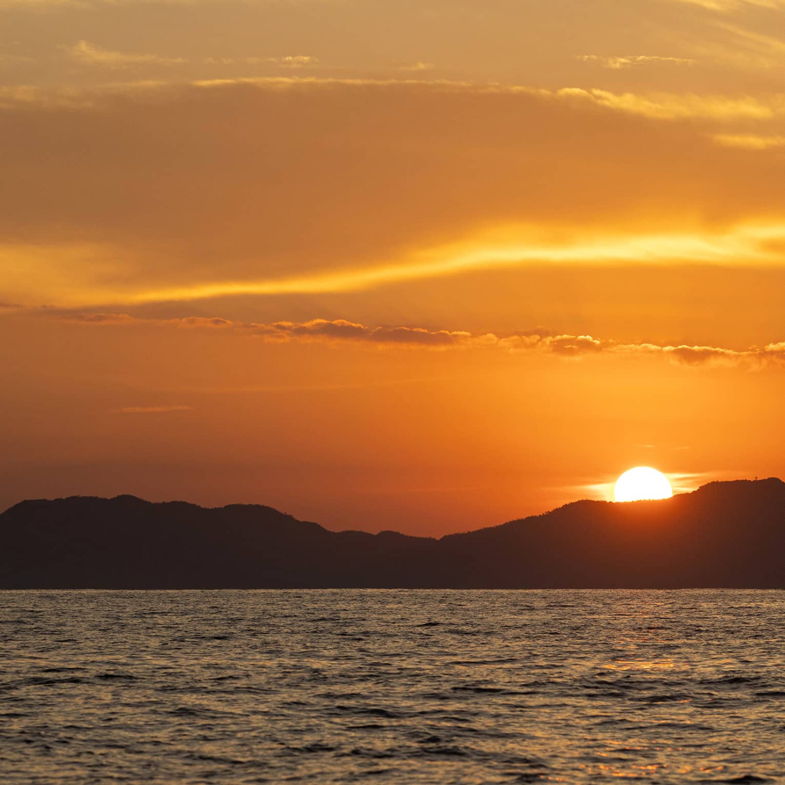 Beyond gently rippling waves, the blazing sun sets behind a hilly island, wispy clouds in the sky awash in its orange glow.