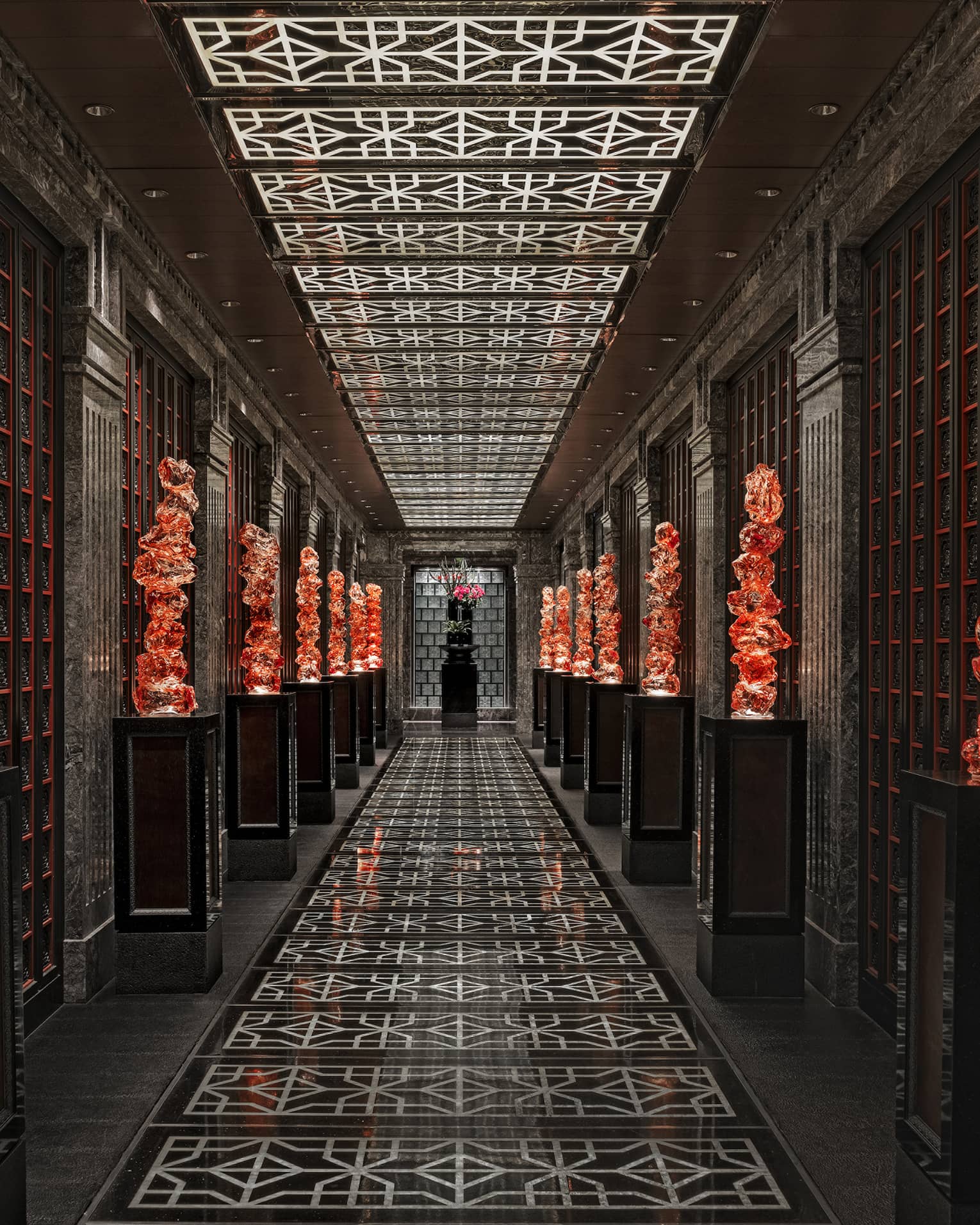 View down a long black-tiled hall lined with orange glass sculptures on pedestals, the geometric ceiling mirroring the floor.