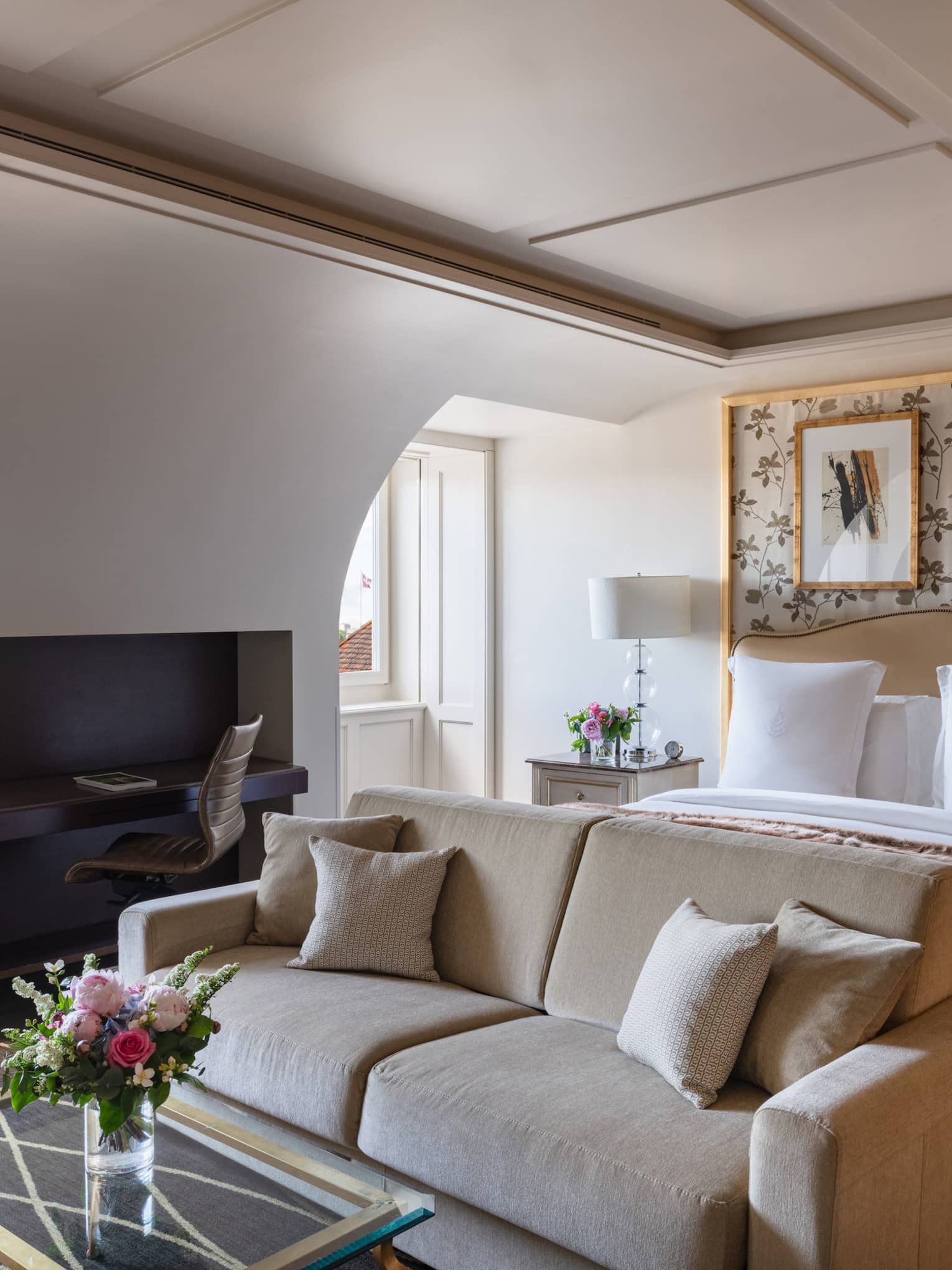 Luxury hotel room with a king bed, beige sofa, coffee table with flowers, built-in desk, floral wallpaper, framed artwork and windows with city view