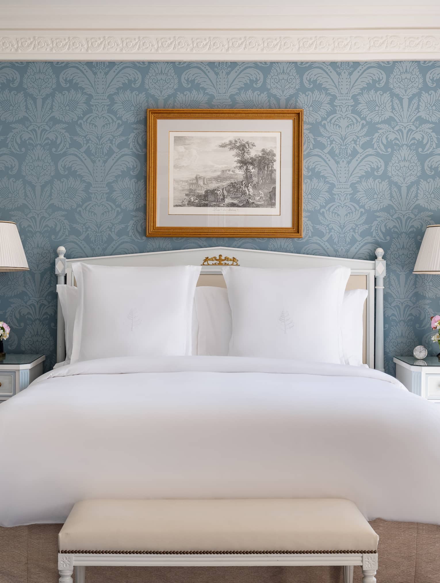 Elegant hotel bedroom with a king bed, white bedside tables with lamps and flowers, bench, blue floral wallpaper and framed artwork