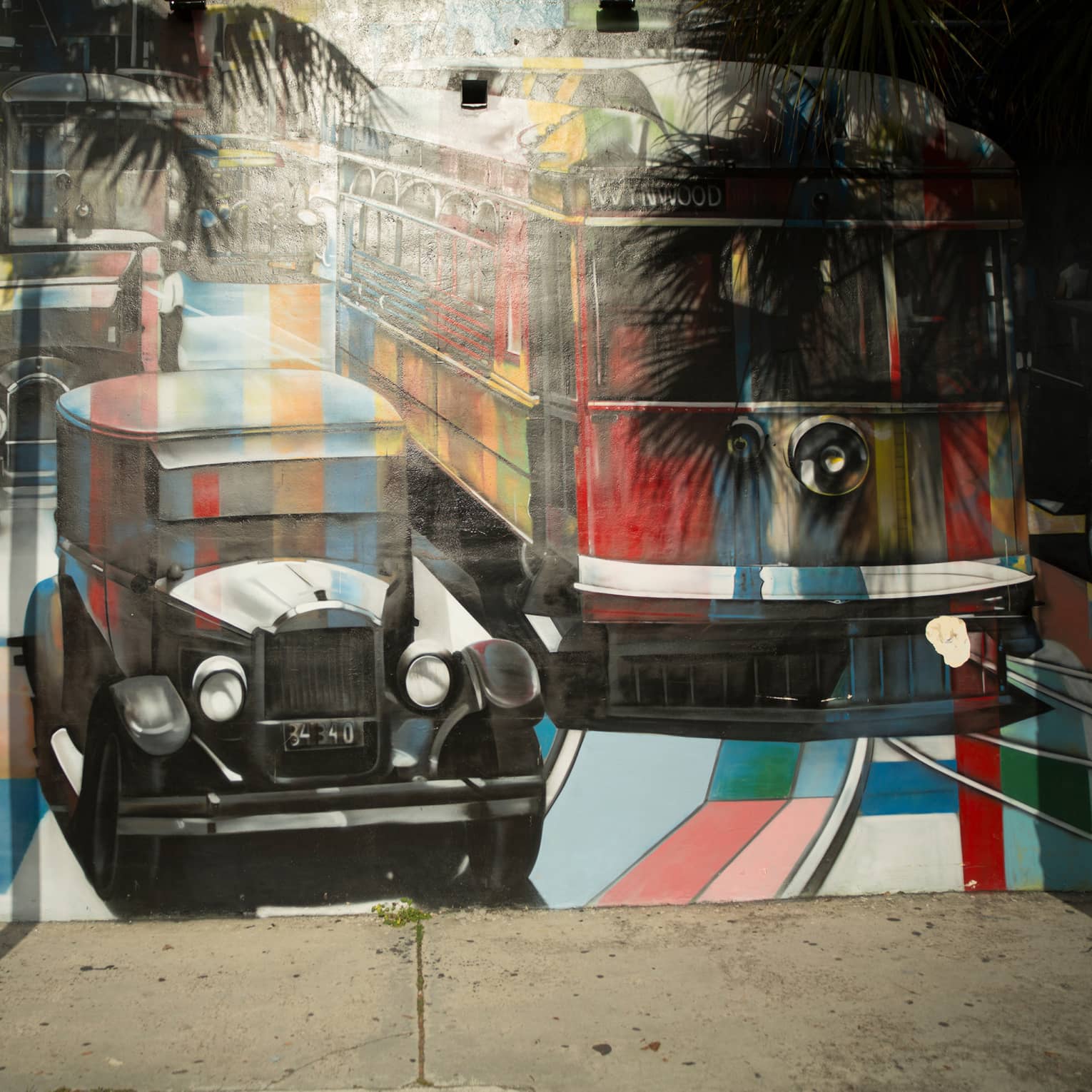 Palm trees cast shadows on a mural of vintage cars and a bus painted in a kaleiscopic pattern of colours and grey shading.
