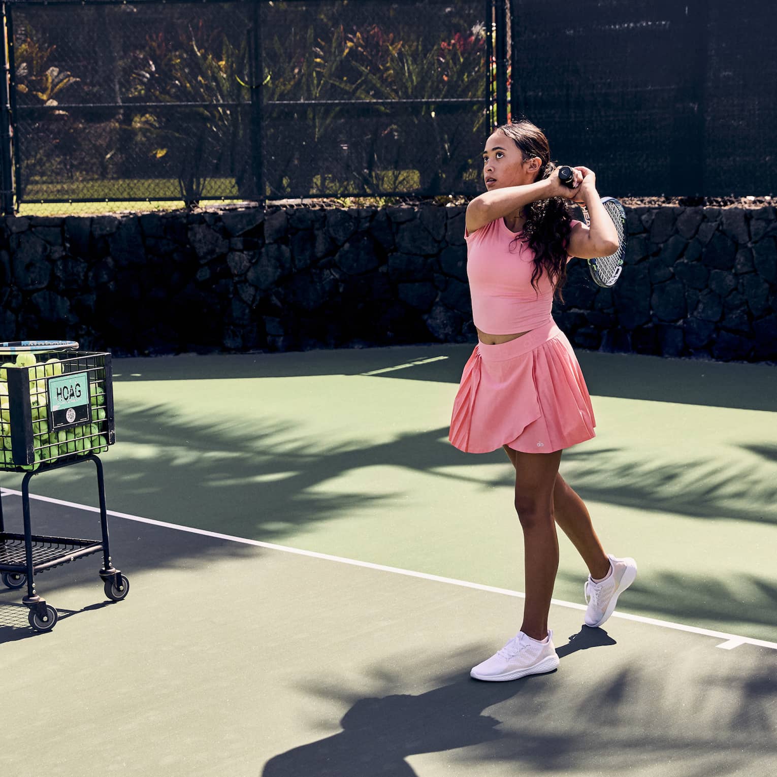 On a tennis court amid palm trees, a player finishes a swing over the shoulder; a pro stands by a ball cart, ball in hand.