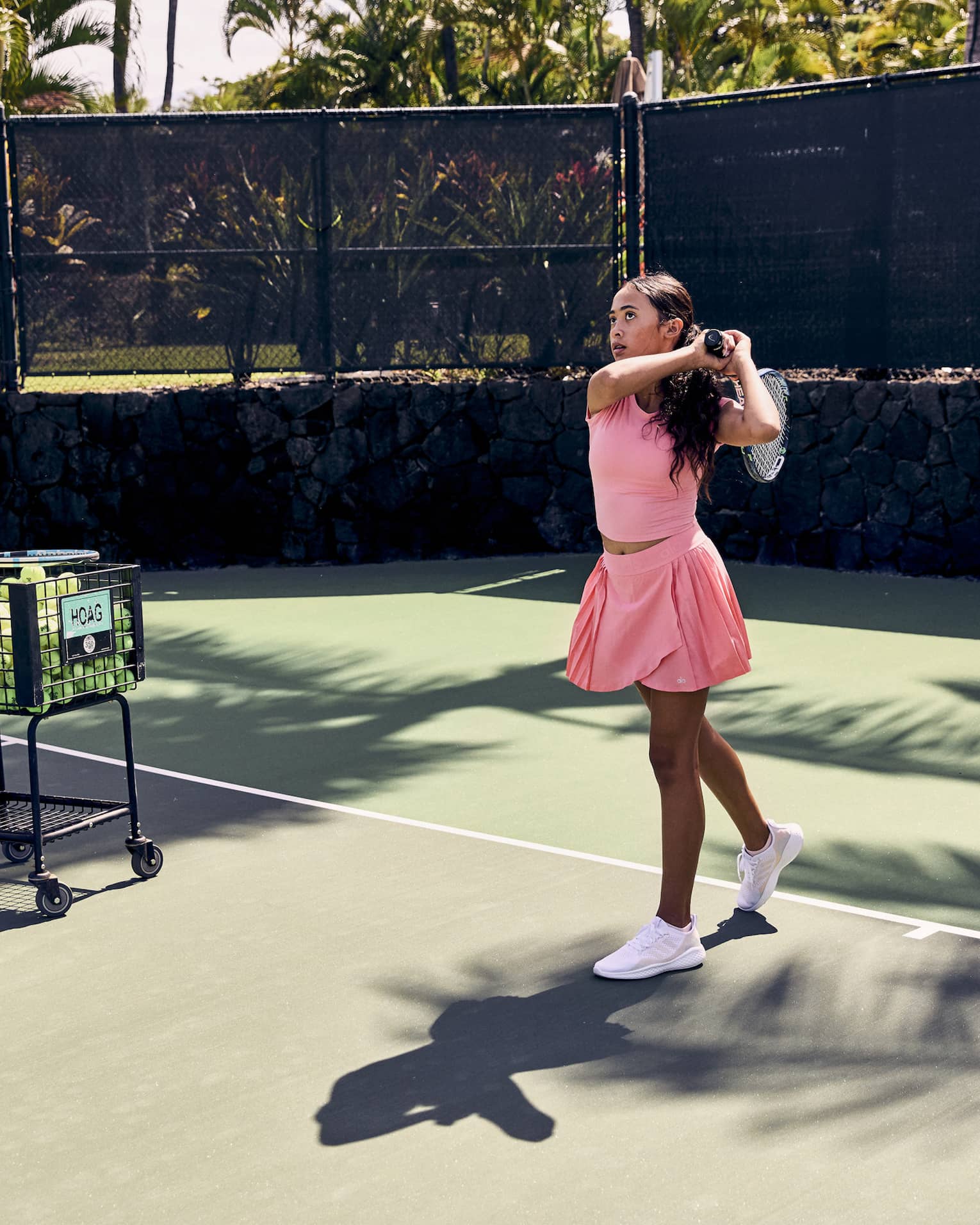 On a tennis court amid palm trees, a player finishes a swing over the shoulder; a pro stands by a ball cart, ball in hand.