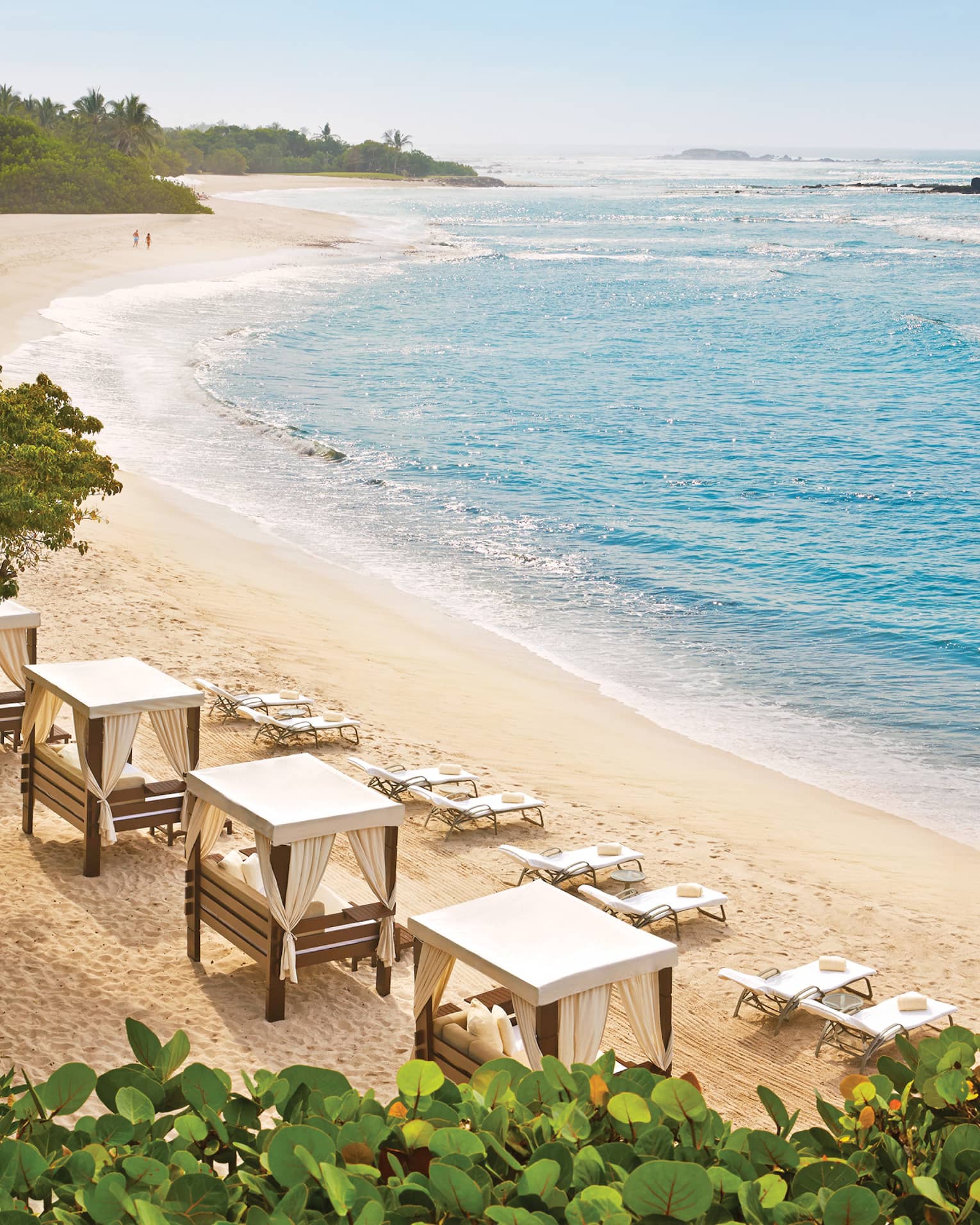 Amid lush vegetation, azure waves roll onto a curved beach lined with chairs and daybeds. A couple strolls in the distance.