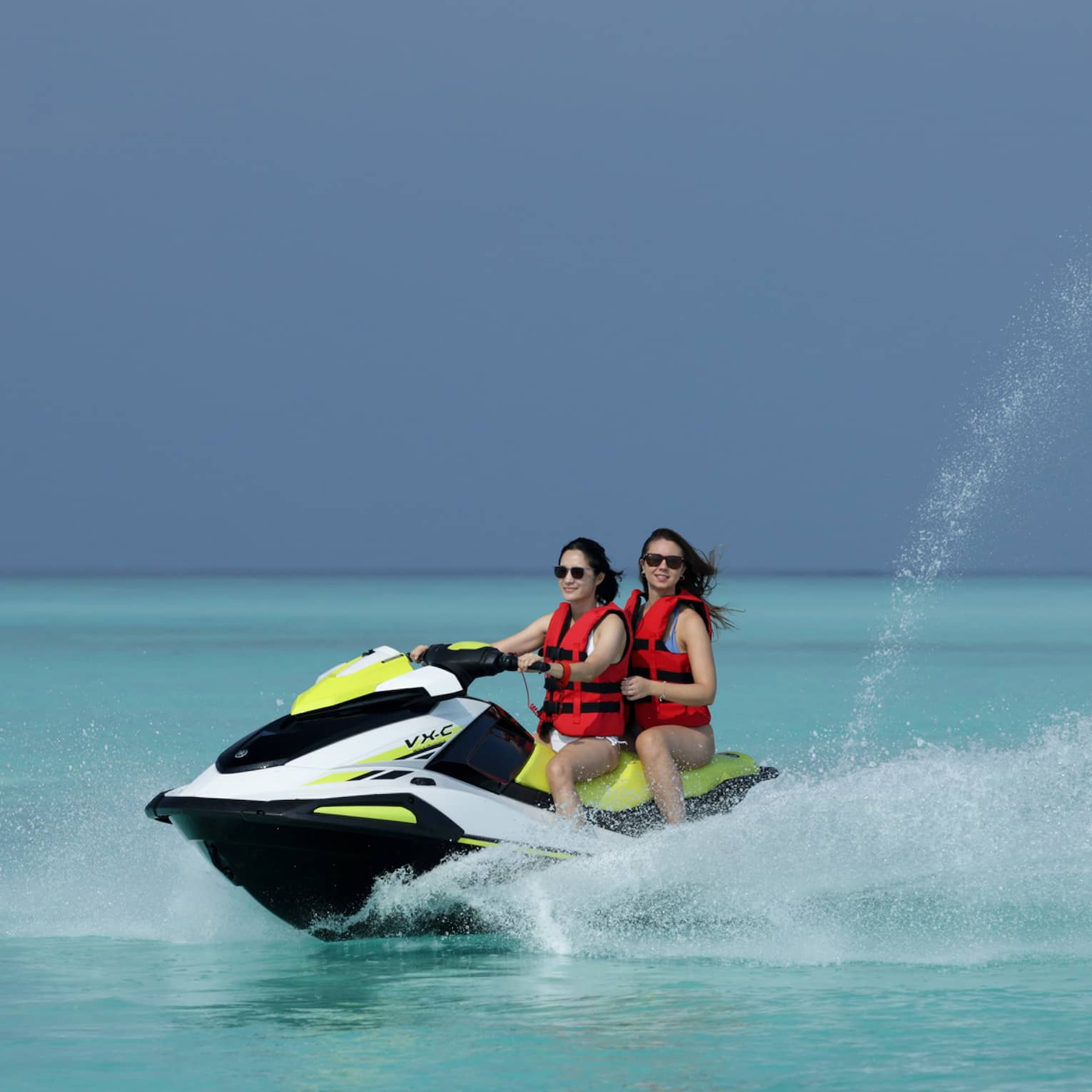 Wearing life jackets, two smiling jet-skiers ride through the clear ocean, water spurting up from beside and behind the boat.