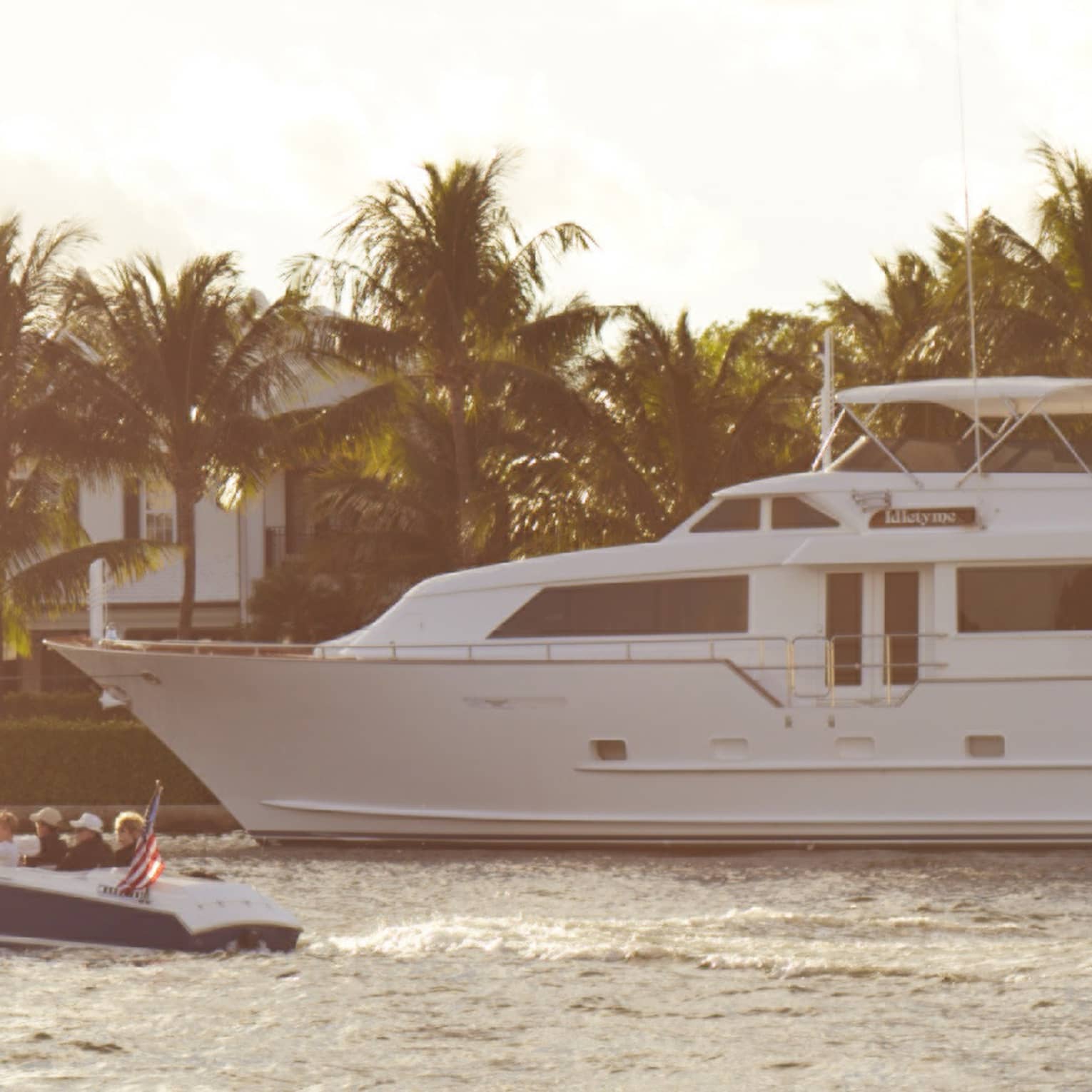 A large white yacht cruises alongside a smaller boat in a serene waterway, with palm trees and luxurious homes in the background under a partly cloudy sky.