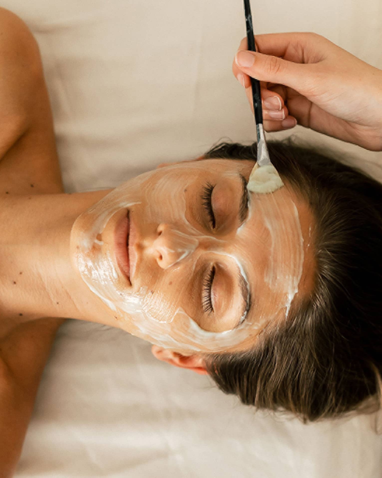 A person holding a small brush applies a white face mask treatment onto a woman's face