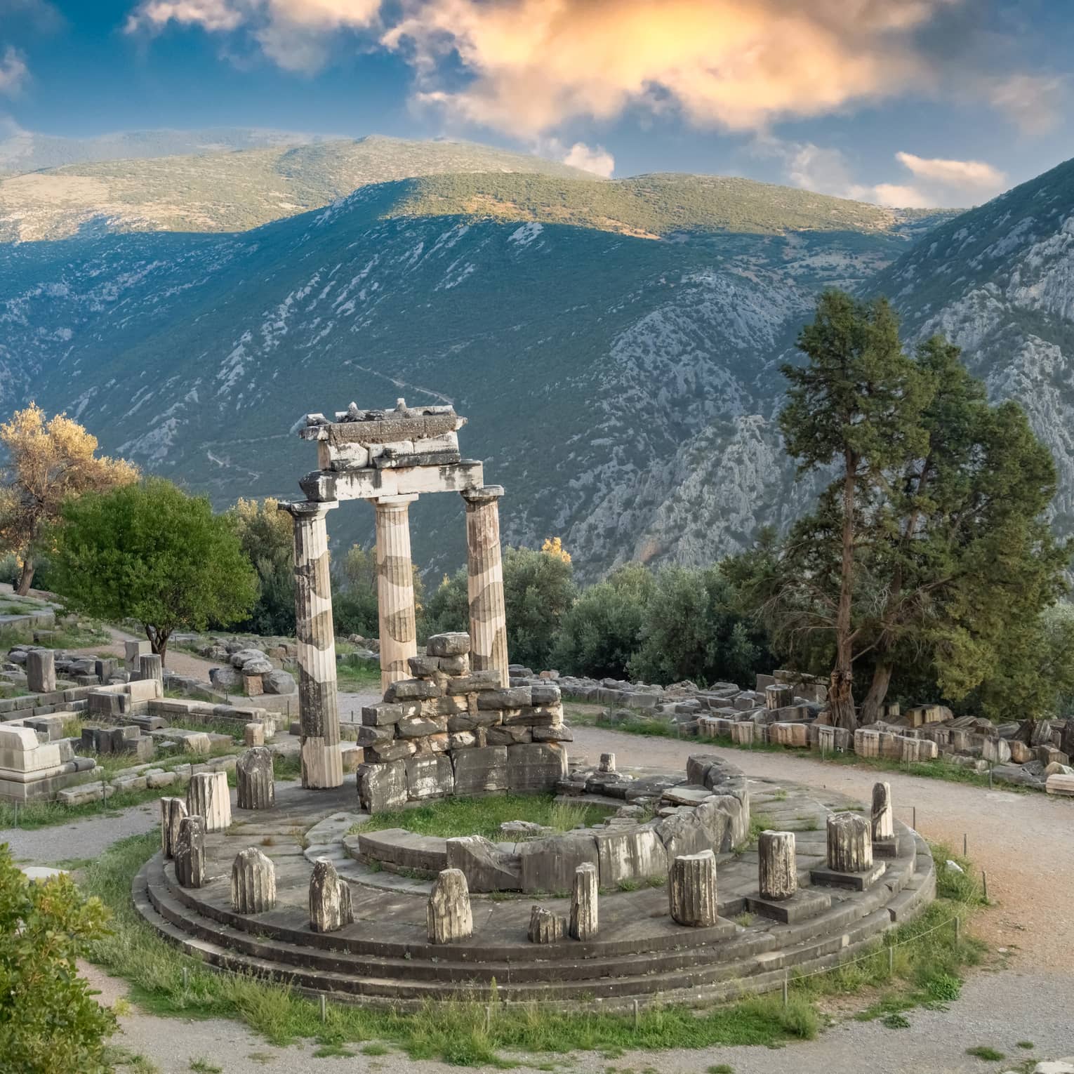 Greek ruins at a hill's edge next to massive green-dappled mountains, under a blue sky with wispy, orange-tinted clouds. 