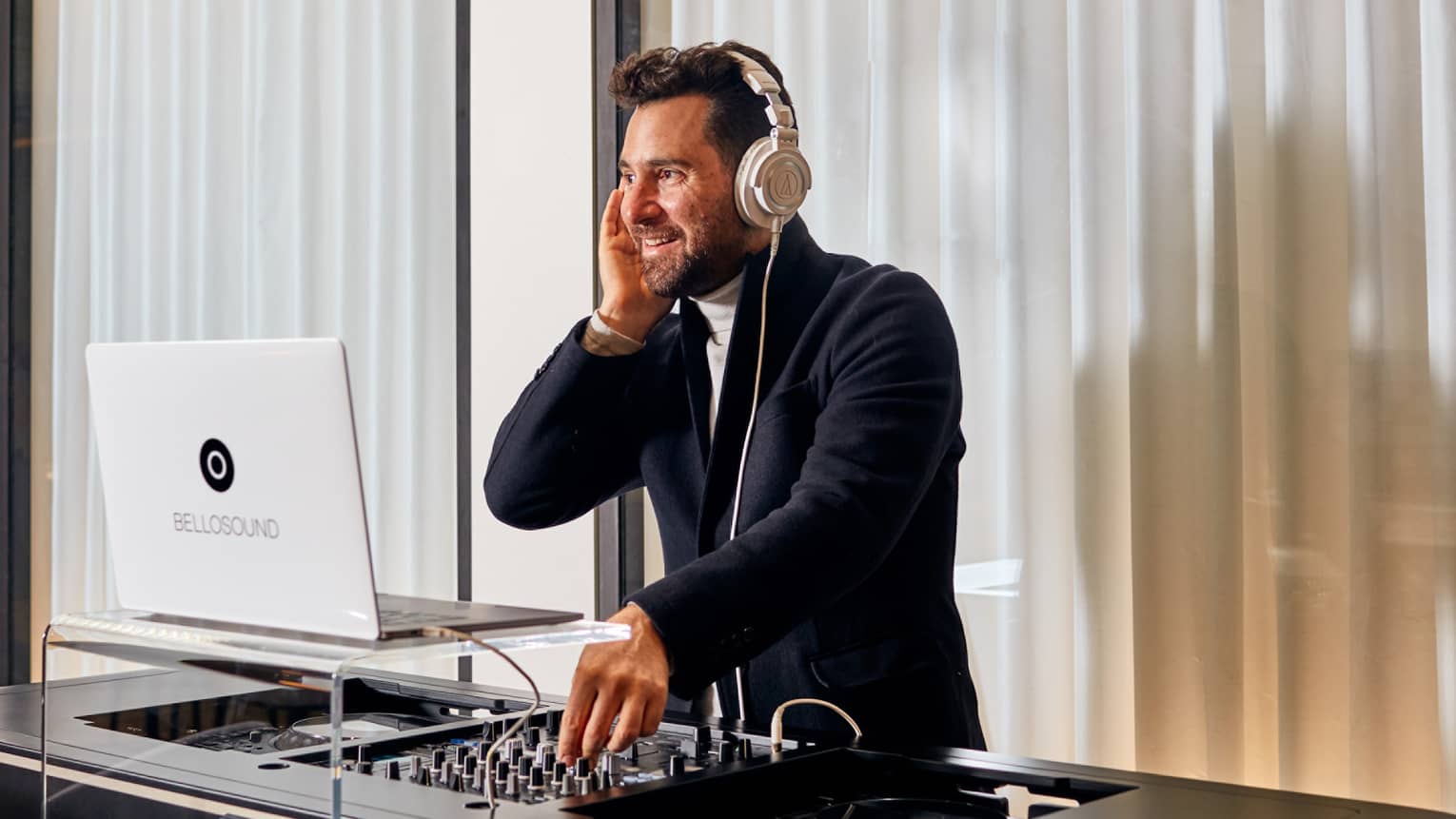 A DJ performing in a white room with closed windows behind him.
