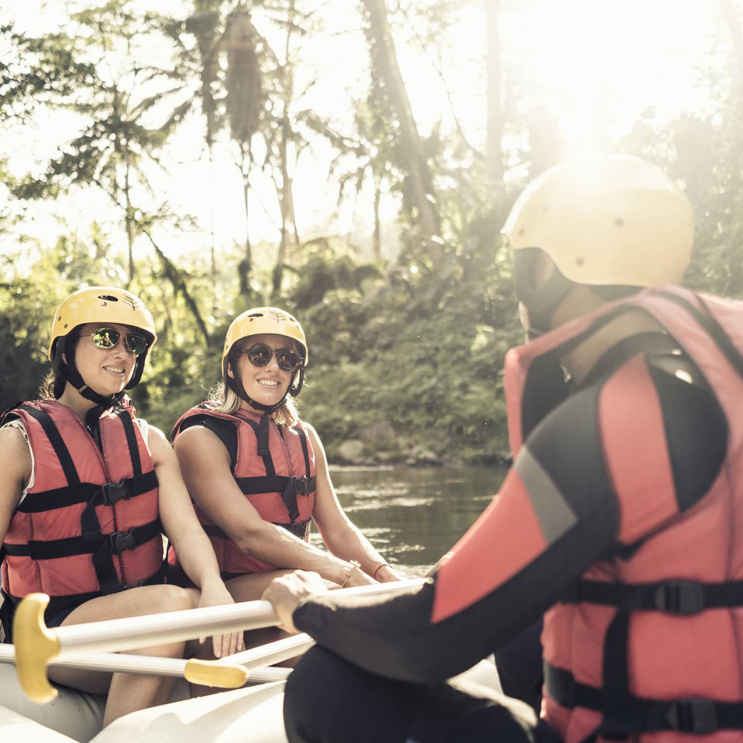 A river rafting guide faces two female guests in the river raft, all wearing red life jackets, sunglasses and yellow helmets with paddles in hand