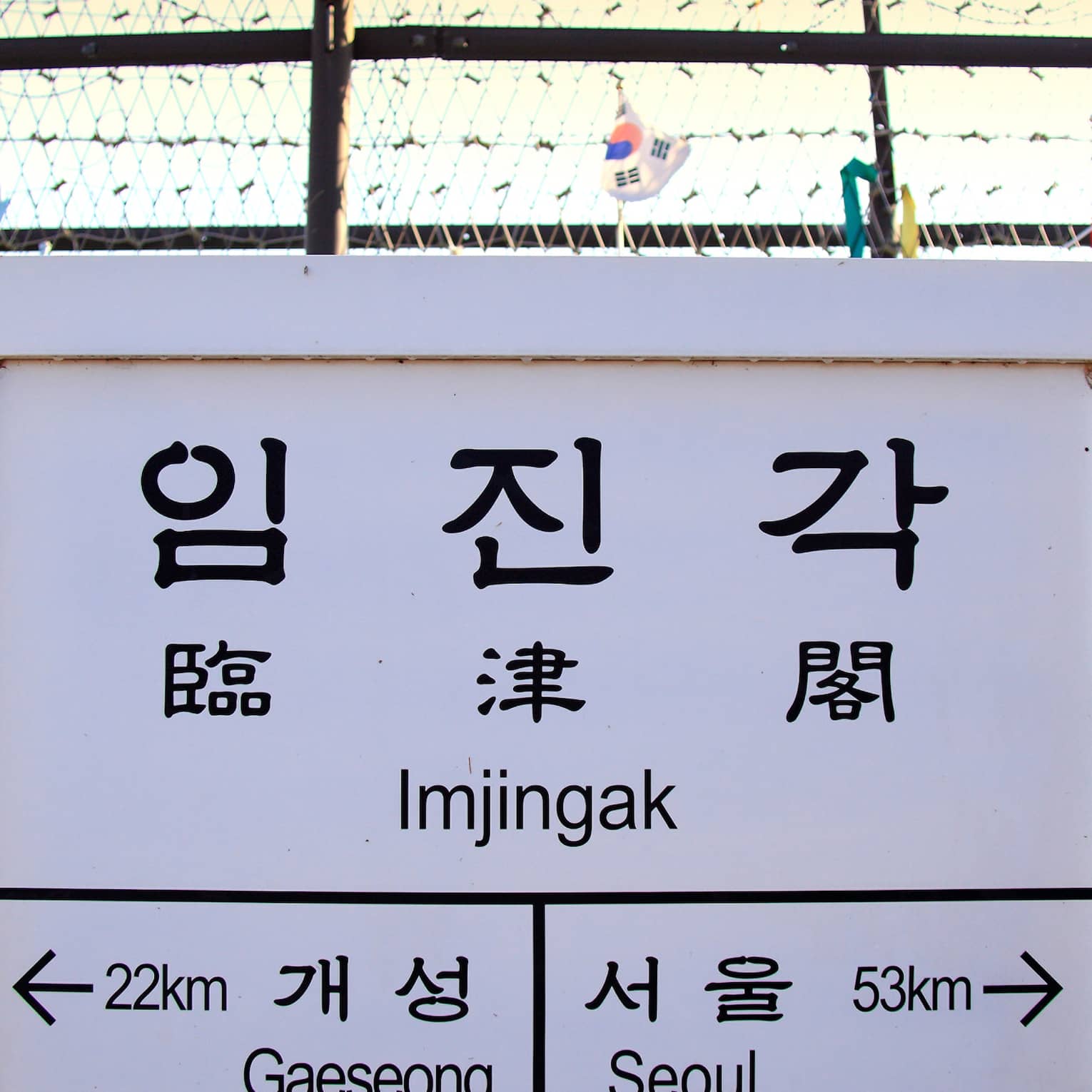 Korean and English text on white sign in front of barbwire fence with ribbons, marking border between North and South Korea.