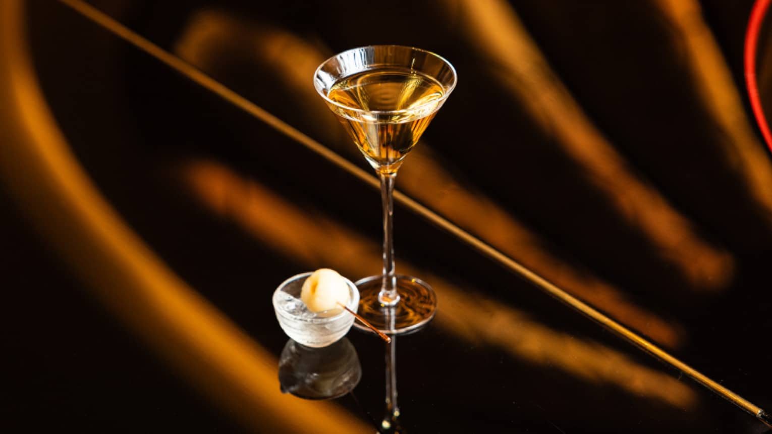 A martini glass on a polished surface holds a pale golden drink.