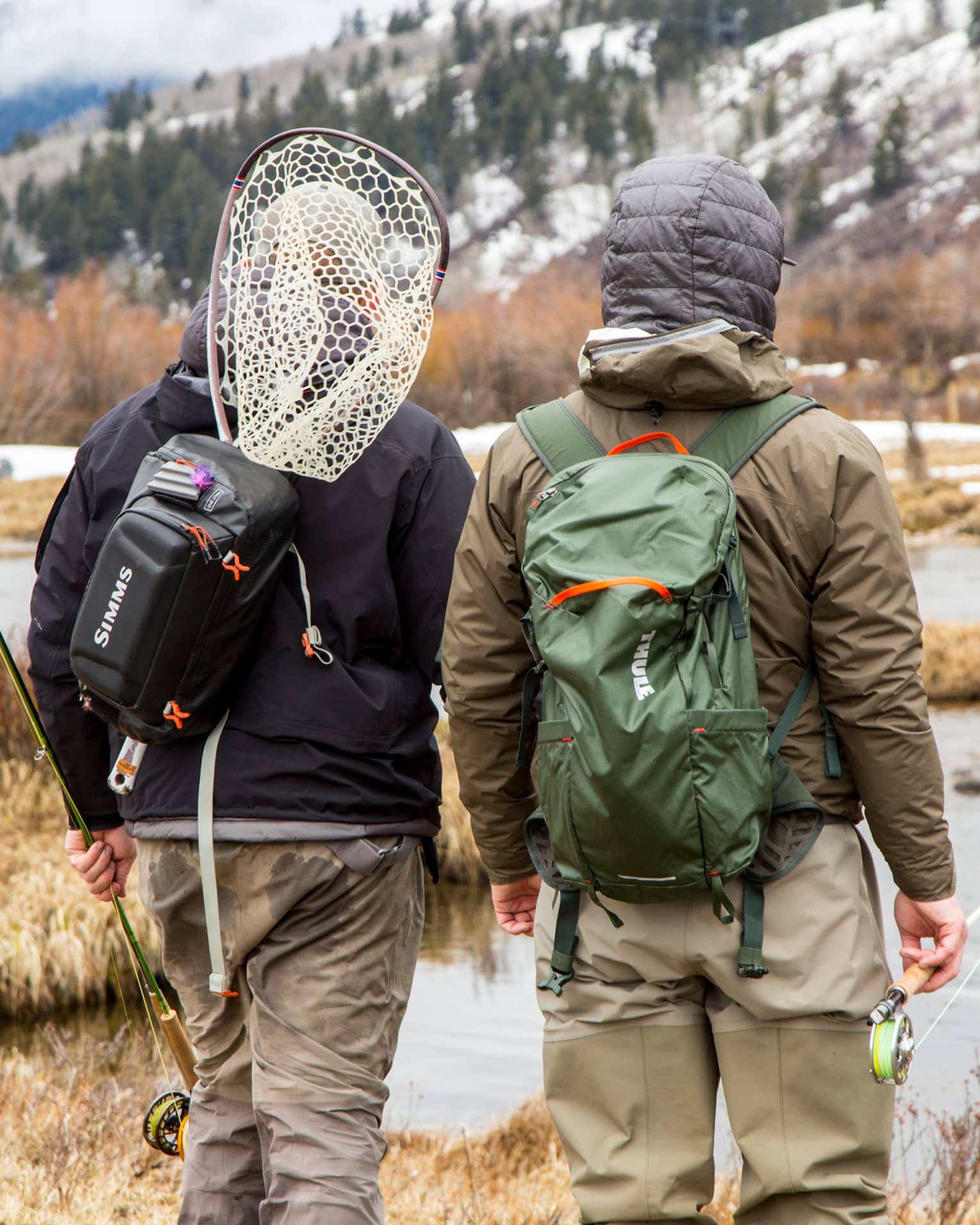 Two people with backpacks and fishing rods look at river with snowy mountain in background 