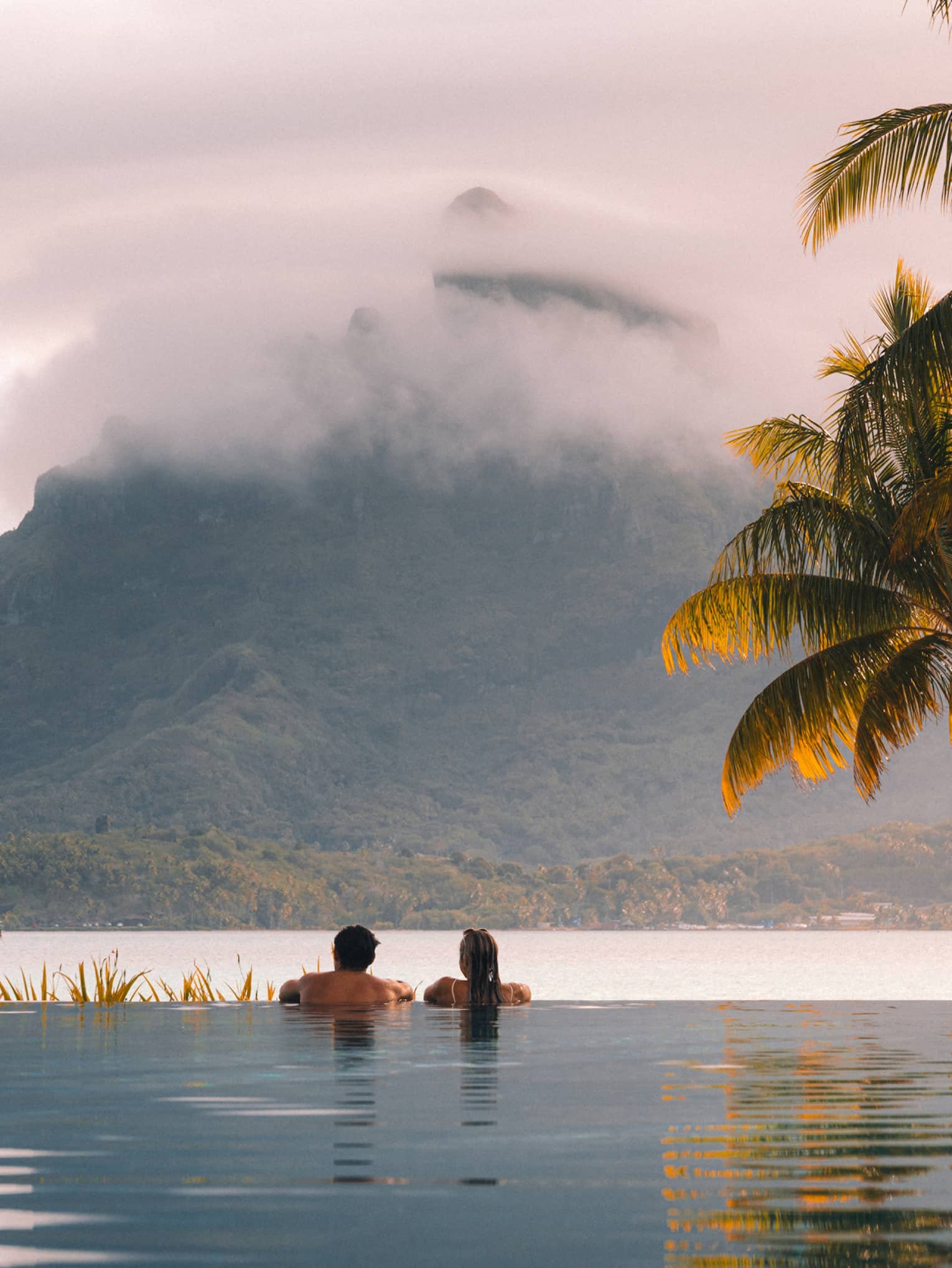 A couple in an infinity pool look across a river at a large mountain in the background. Palm trees and grasses line the pool.