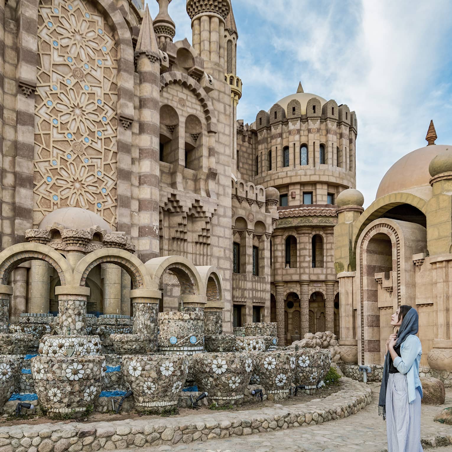 A guest stands in front of a large, intricately tiled stone building, gazing at its numerous turrets, archways and domes.