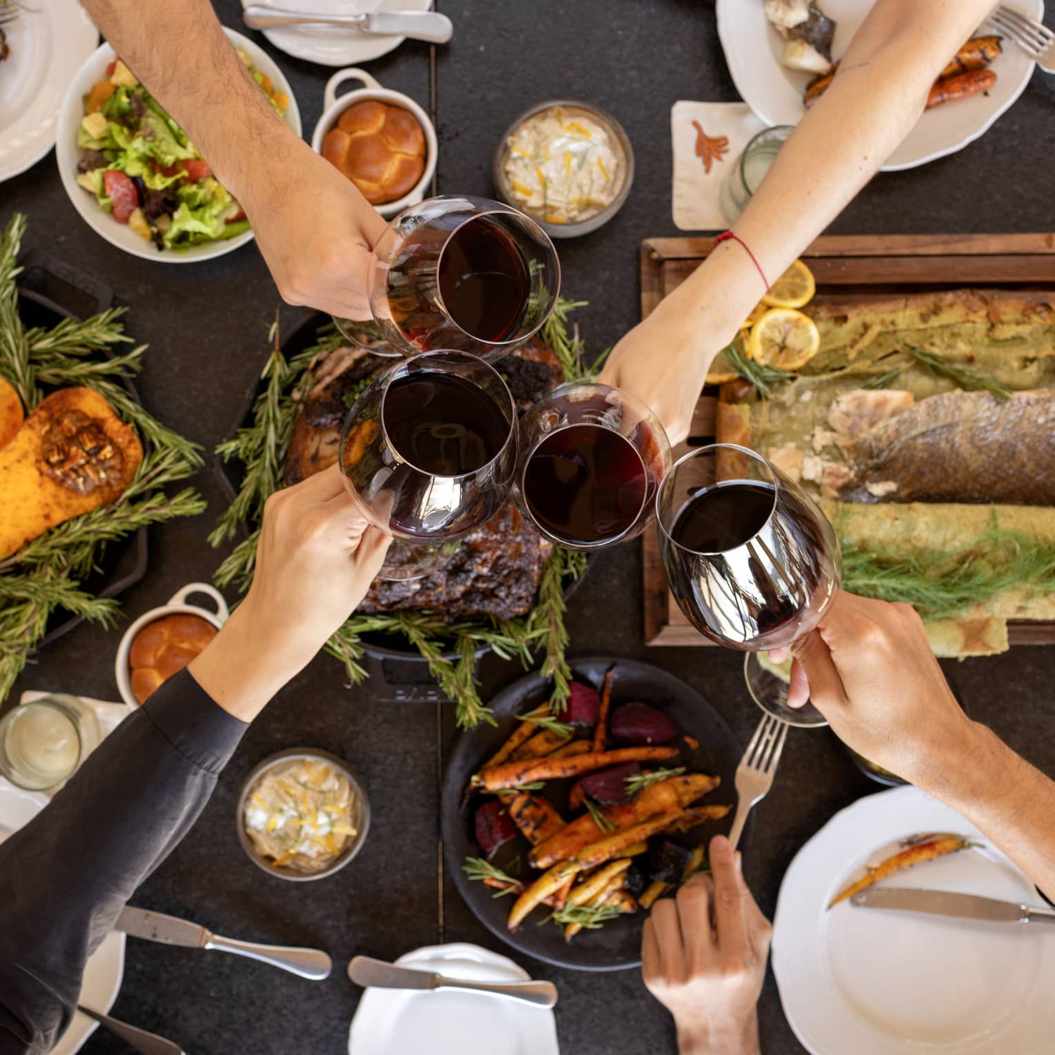 Overhead view of a table filled with festive dishes such as roasted squash, a filet of fish and other platters with four hands each holding a glass of red wine coming together in the centre