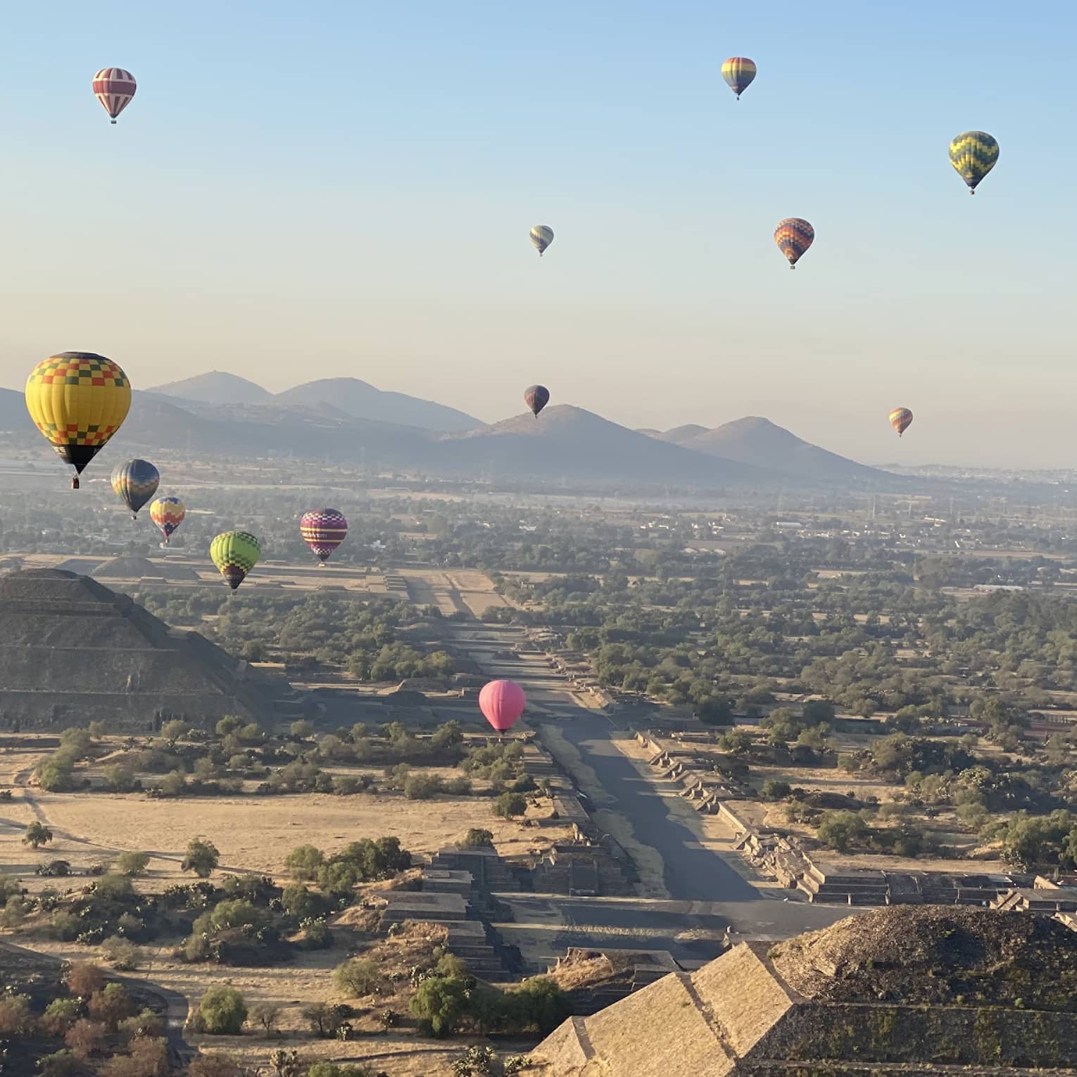 An aerial view of colourful hot-air balloons floating over pyramids and trees, with mountains and a city in the background.