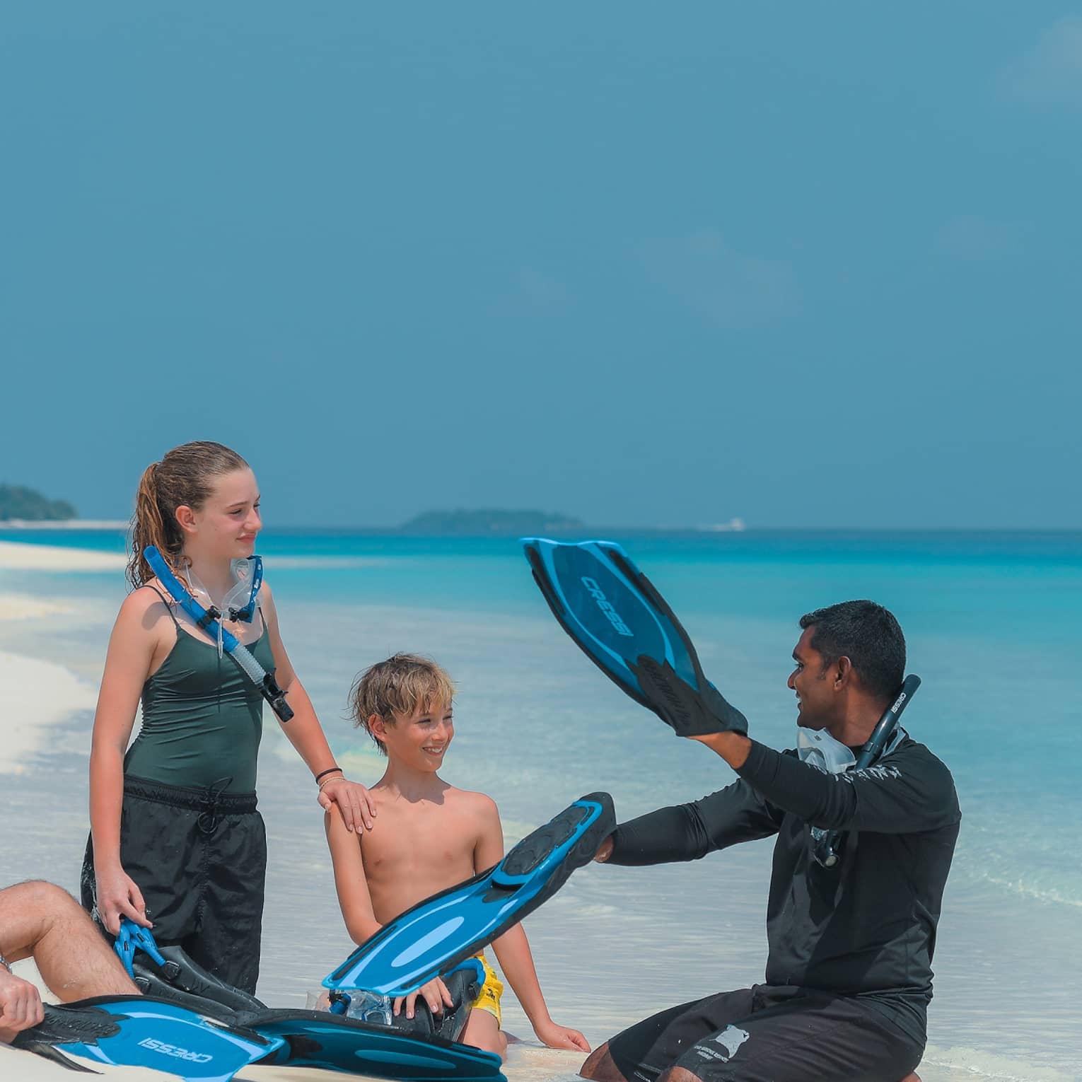 Sitting on a white sandy beach by the water, a family with snorkel gear intently listens to the instructor holding flippers.