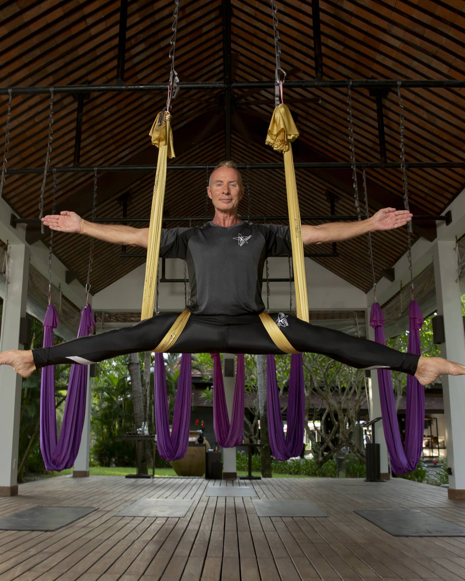 In a covered gazebo, the instructor smiles serenely while suspended in a gold silk sling, arms and legs outstretched..