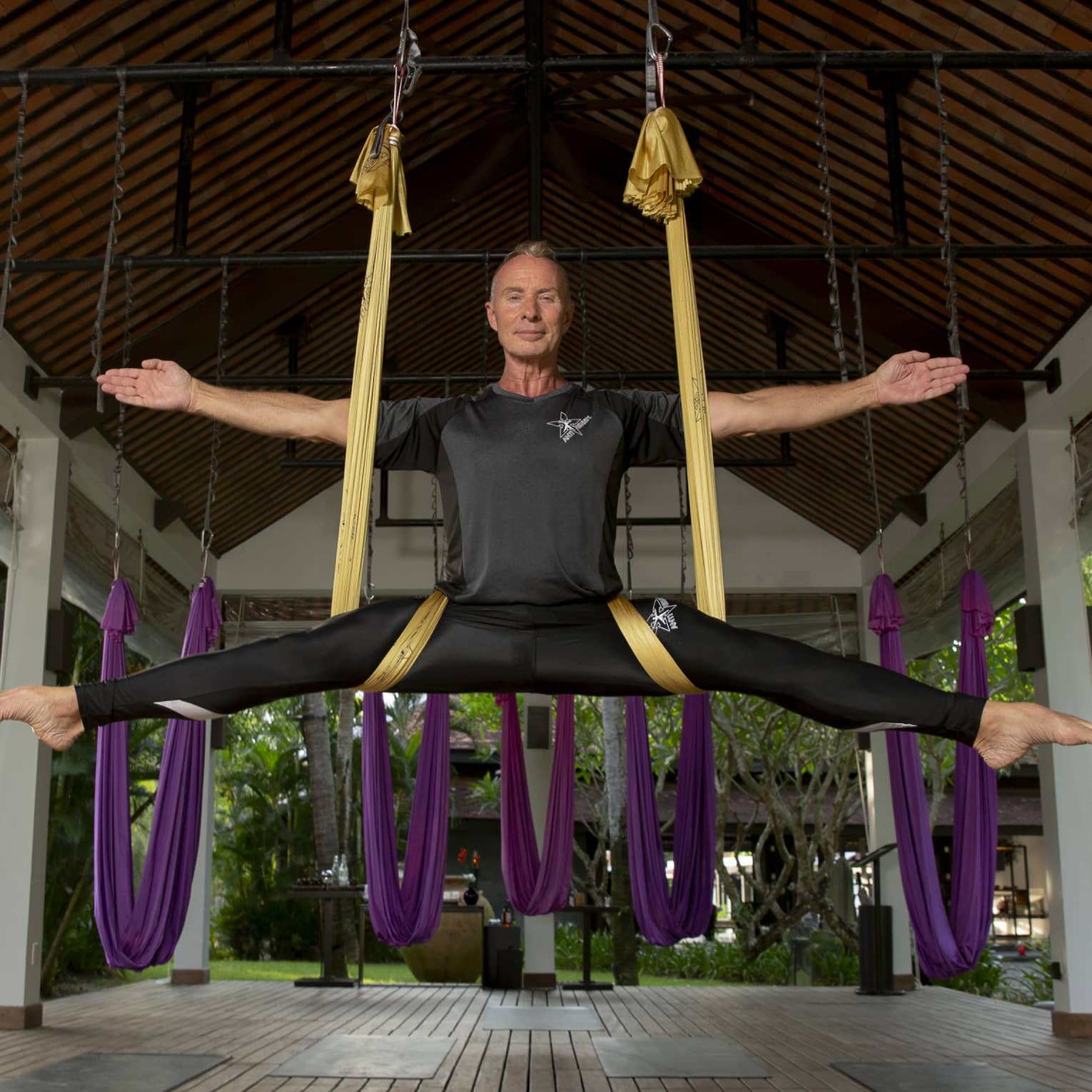 In a covered gazebo, the instructor smiles serenely while suspended in a gold silk sling, arms and legs outstretched..