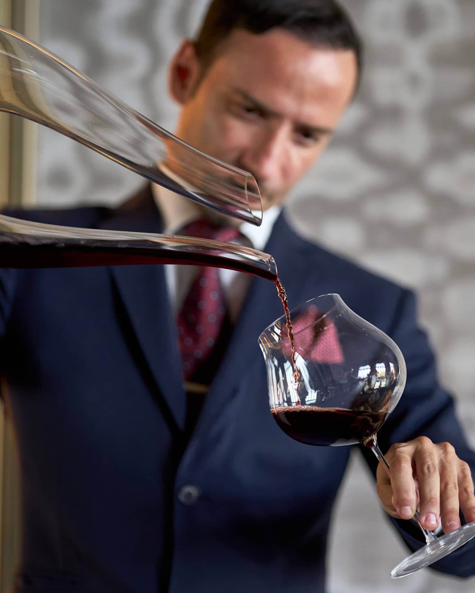 A person in formal attire carefully pouring red wine from a decanter into a glass.