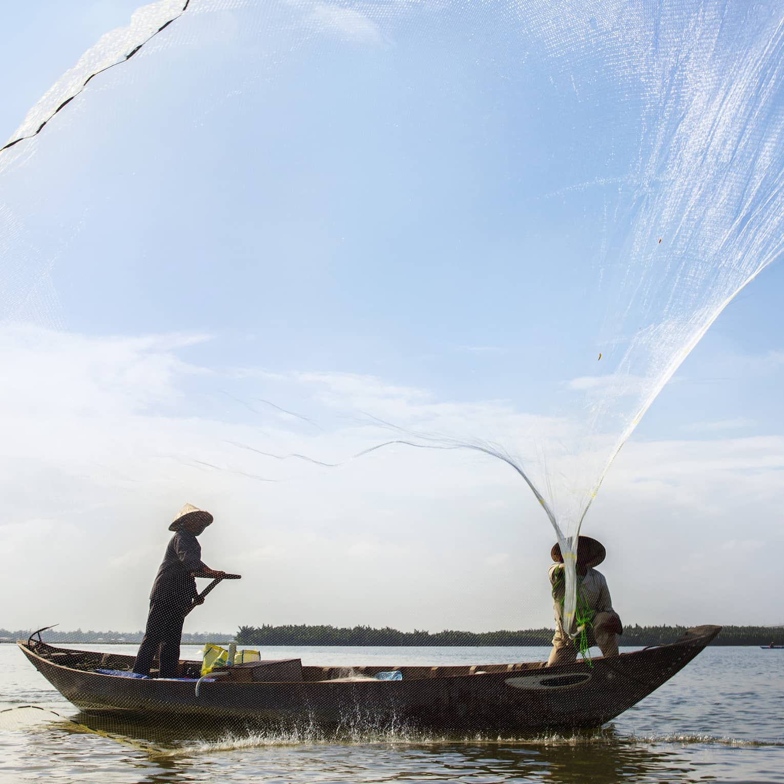Silhouettes of two people wearing hats in small boat standing, casting large nets 