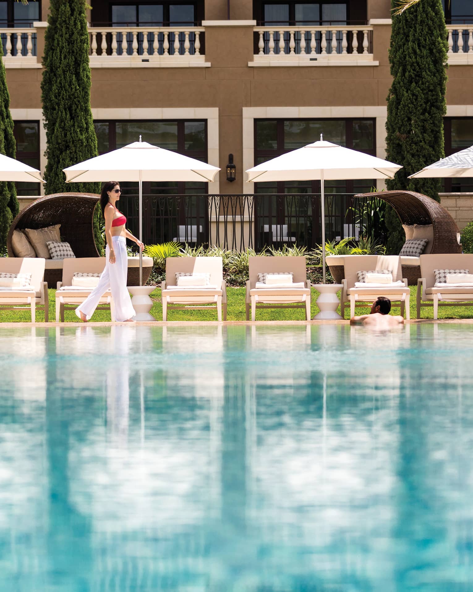 Woman walks by row of white patio chairs, umbrellas on outdoor swimming pool deck