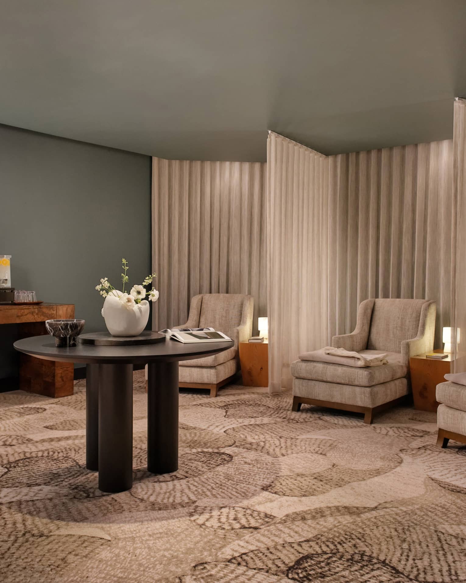 Spa relaxation area with tan armchairs separated by curtains, round table in middle of room with vase of flowers