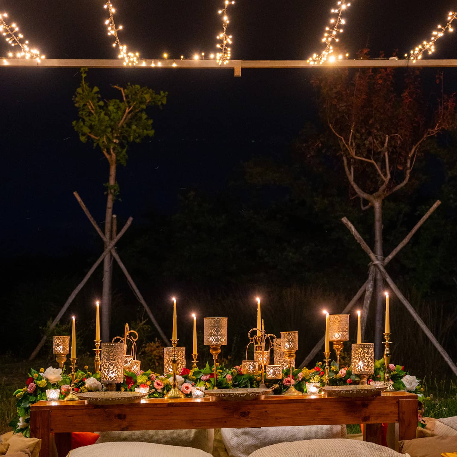 Low table with lit taper candles and gold lanterns, lush floral runner, surrounded by large cushions, under string lights.