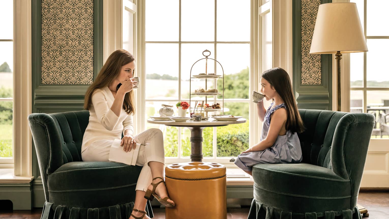 Woman and young girl sit in velvet armchairs by sunny window, enjoy afternoon tea service