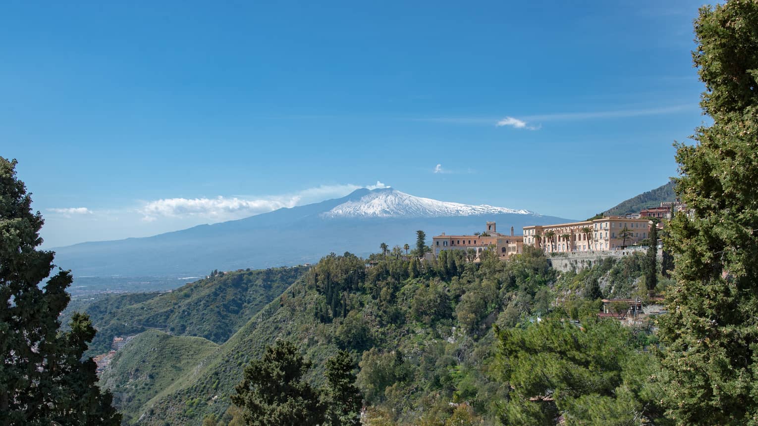 View from afar of an expansive mediterranean villa atop a forested hill, snow-capped mountain in the distance.   