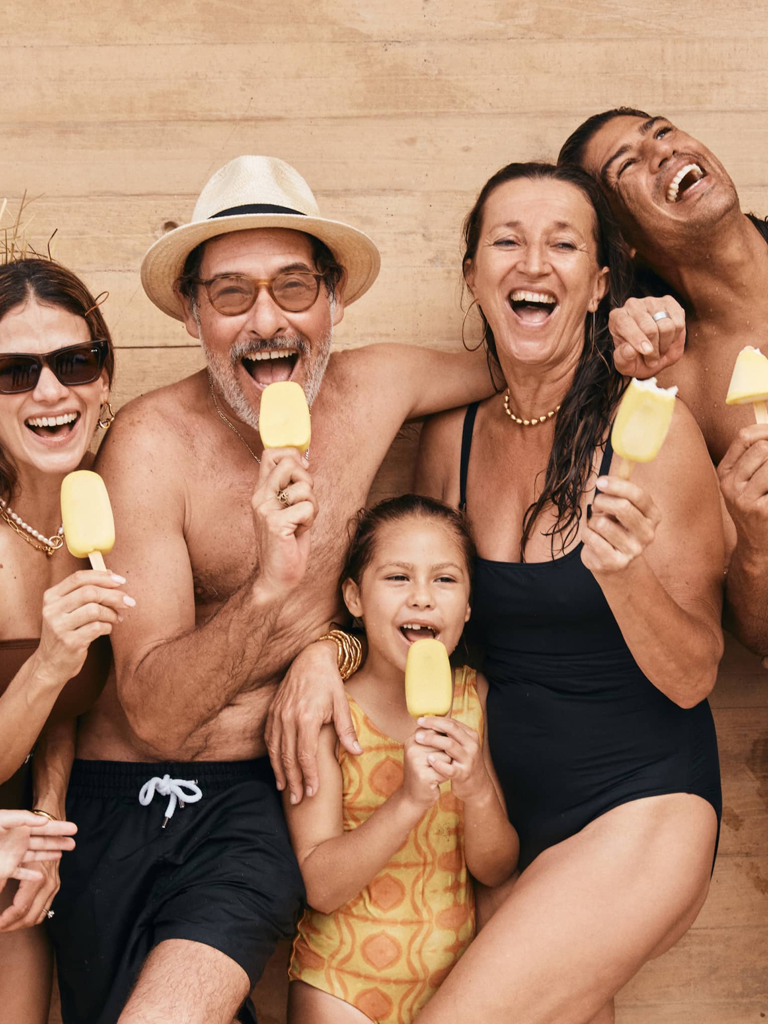 A group of adults and children in bathing suits hold yellow popsicles and smile at the camera.
