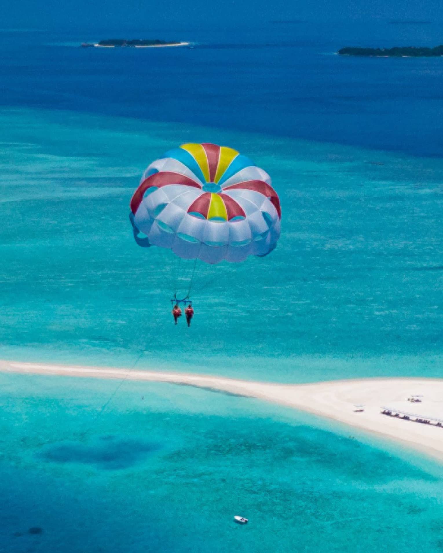 Aerial view of two people parasailing in tandem, colourful sail floating above an expanse of aqua water and a white sandbar.