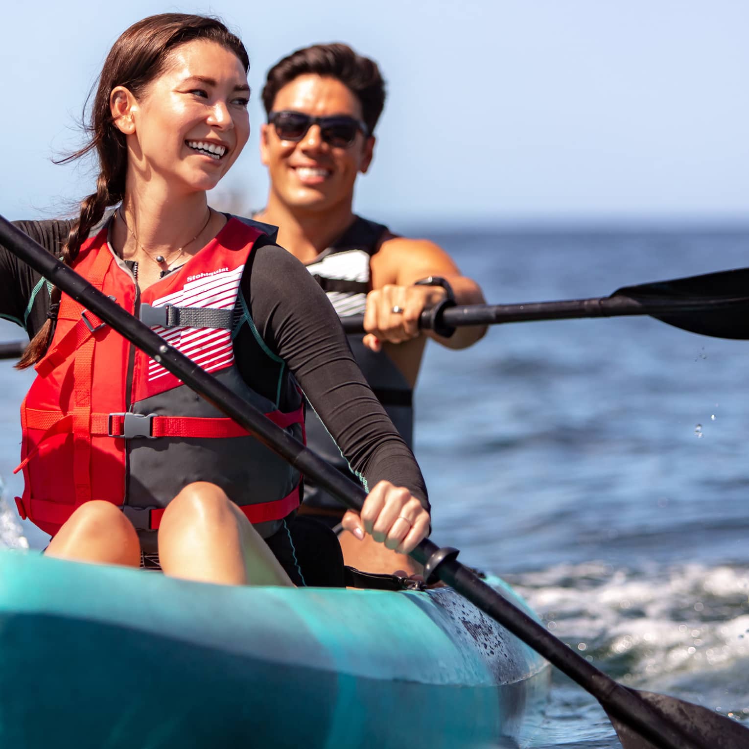 Close-up front view of two smiling adults paddling in a tandem kayak on open water under a sunny blue sky.