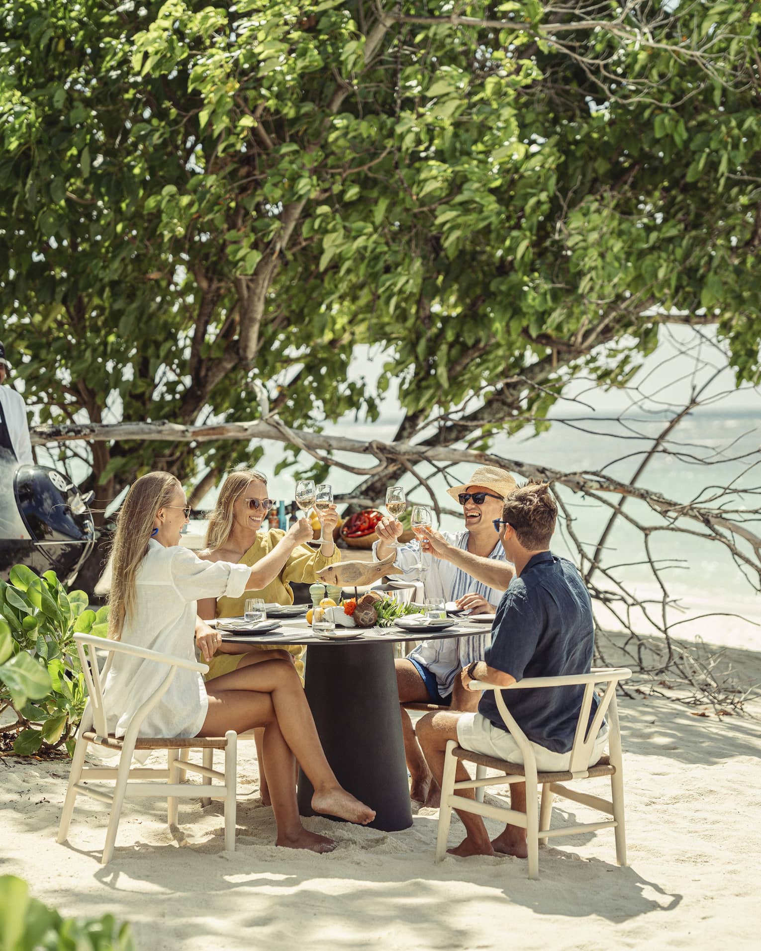 A group sits at a round table for a beach-side meal, clinking glasses while a smiling chef cooks on a coal barbecue nearby. 