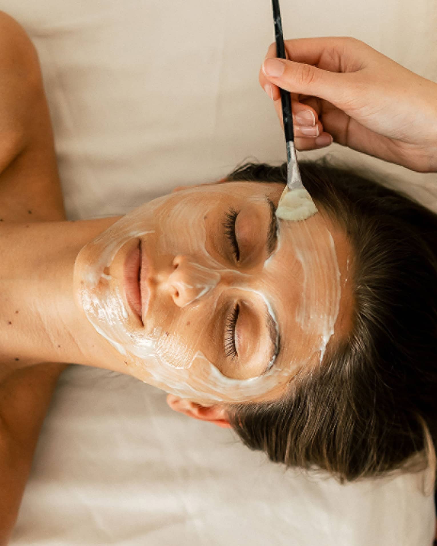 A person holding a small brush applies a white face mask treatment onto a woman's face