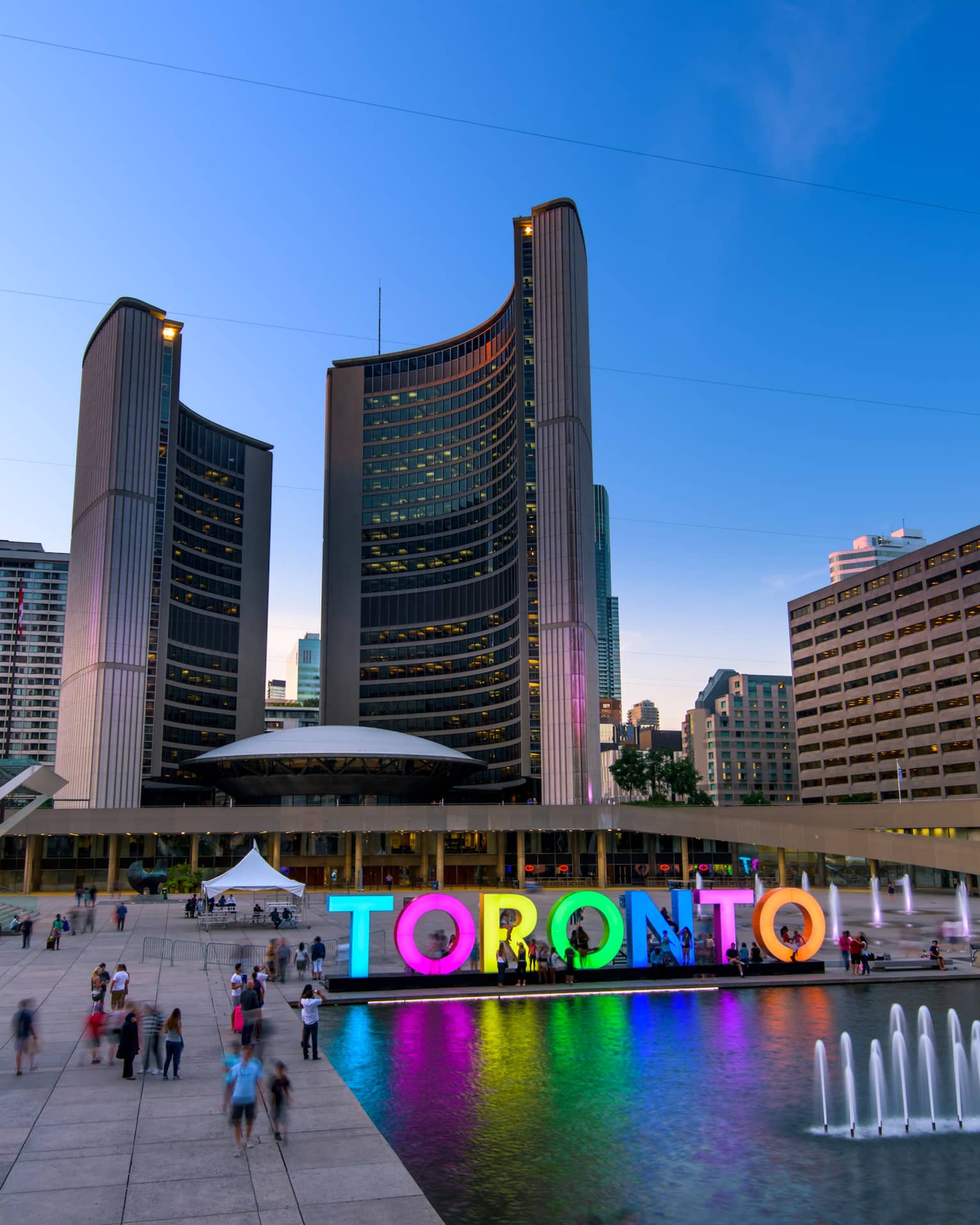 Colourful glowing letters spelling Toronto by fountain people in courtyard under curved City Hall buildings